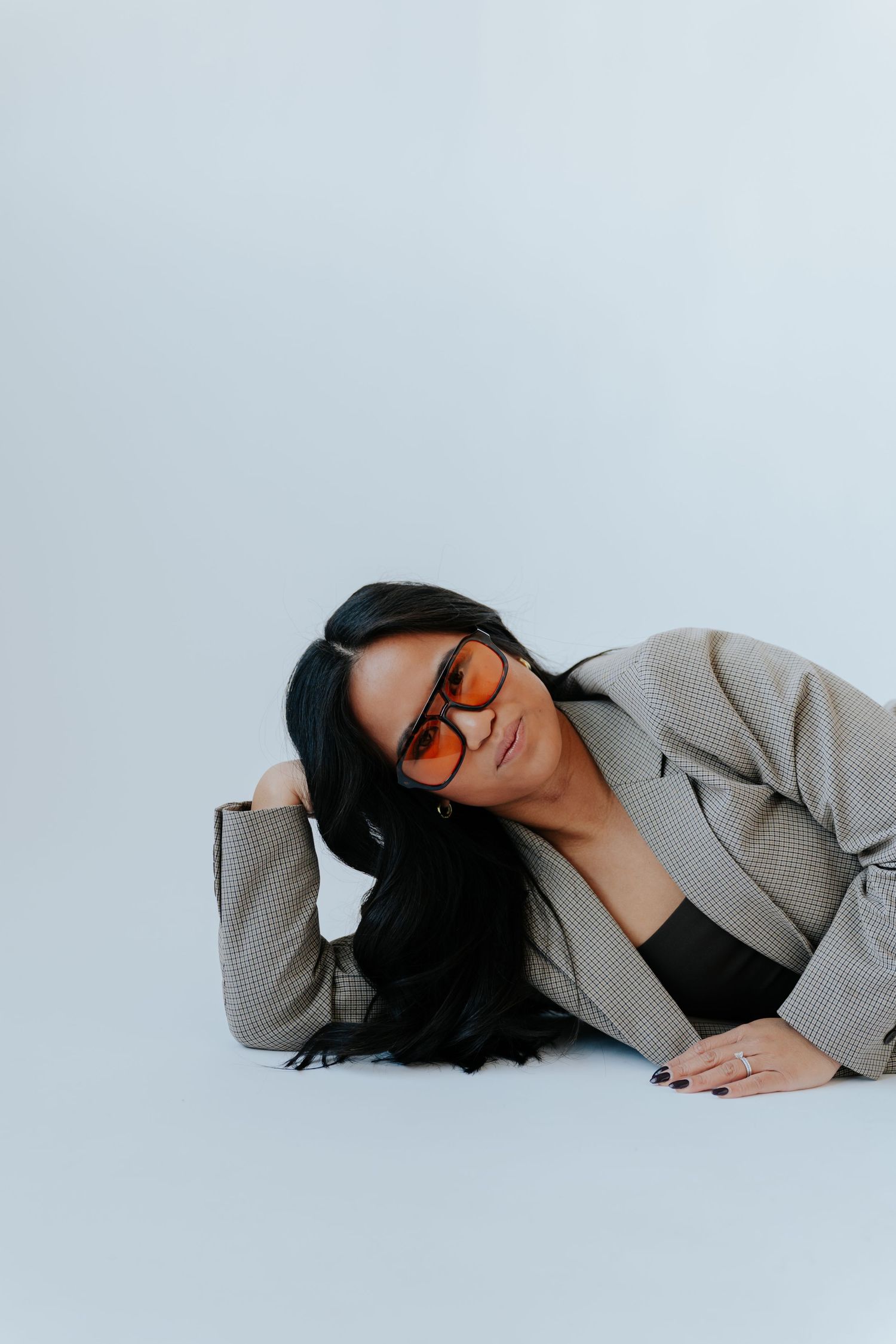 Woman in plaid blazer and orange sunglasses posing on floor against light gray background.