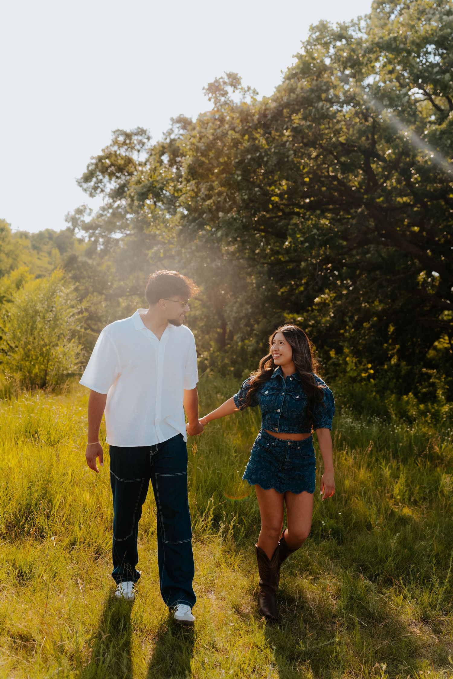 Couple holding hands walking through a sunlit green meadow, woman in teal denim outfit and boots, man in white shirt and jeans.