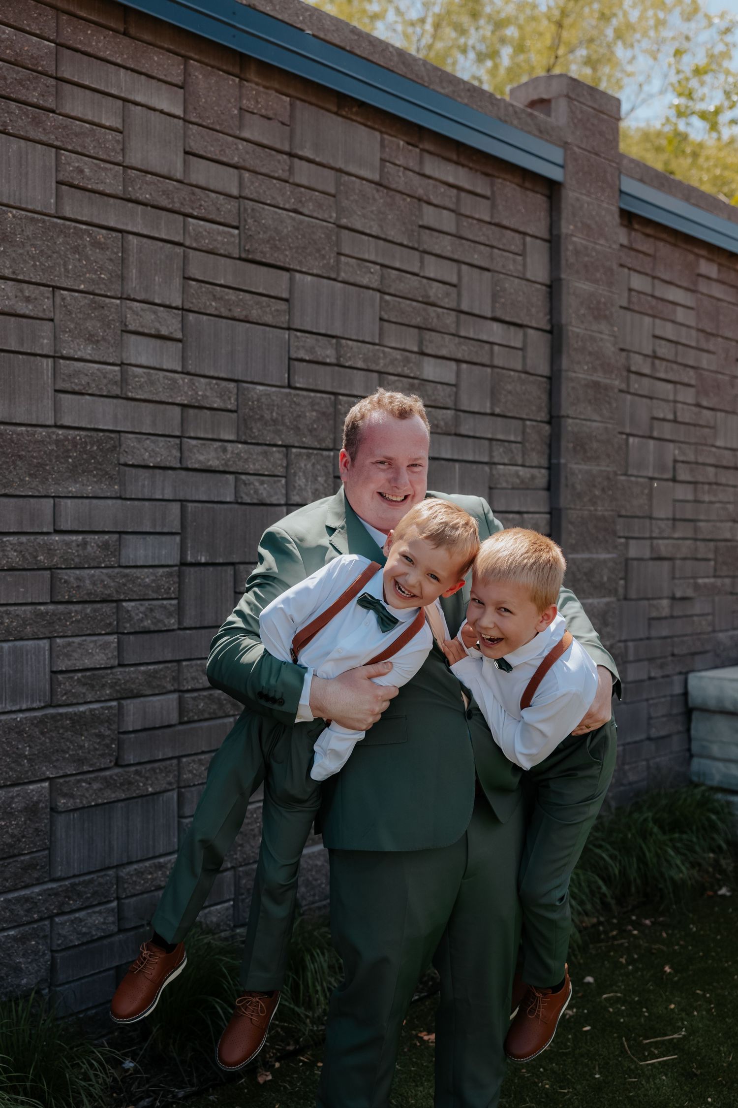 Smiling groom in green suit hugging two young boys in suspenders against a stone brick wall outdoors.