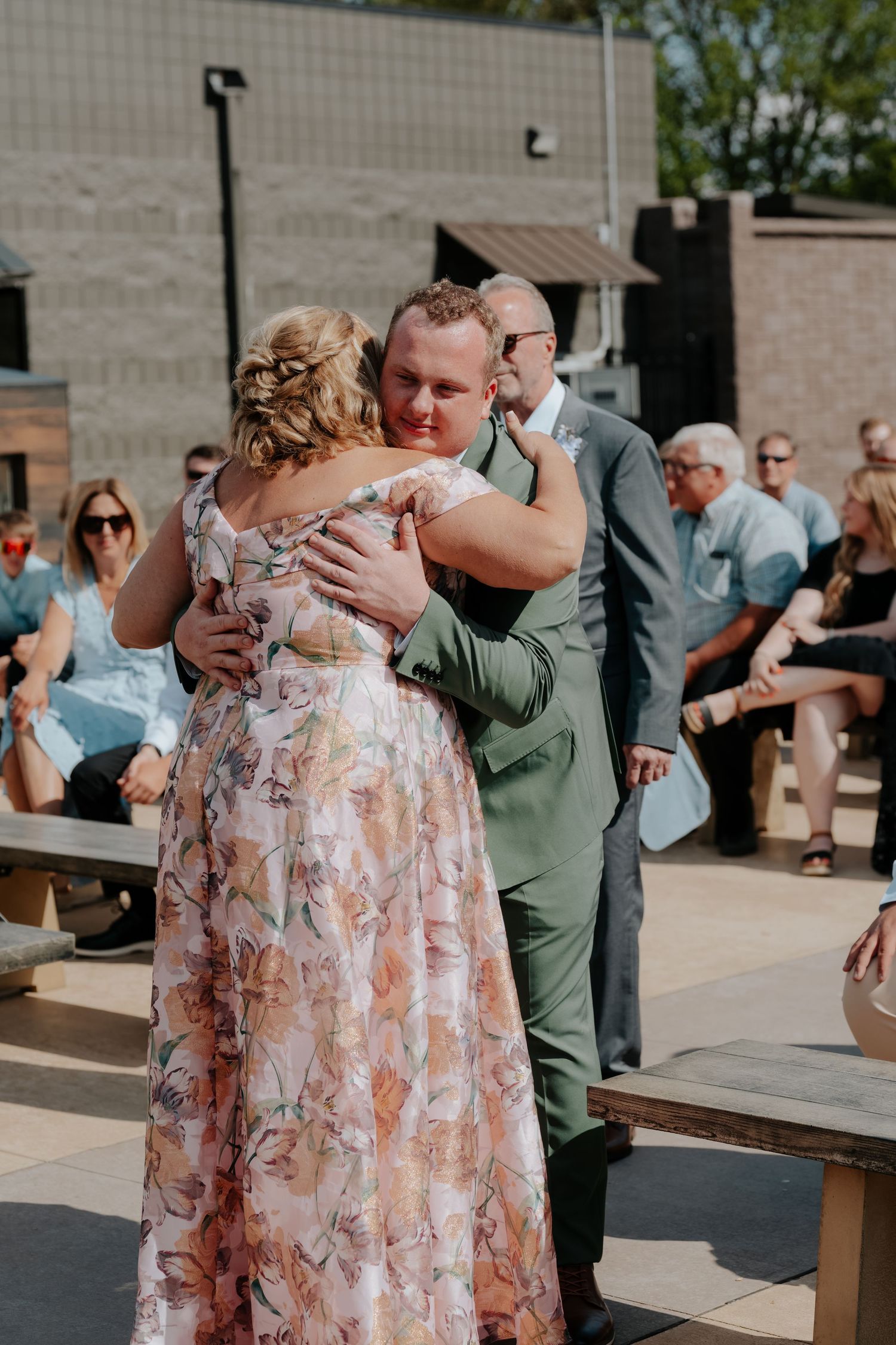 Groom in green suit hugging a woman in floral dress at outdoor wedding ceremony with guests seated behind.
