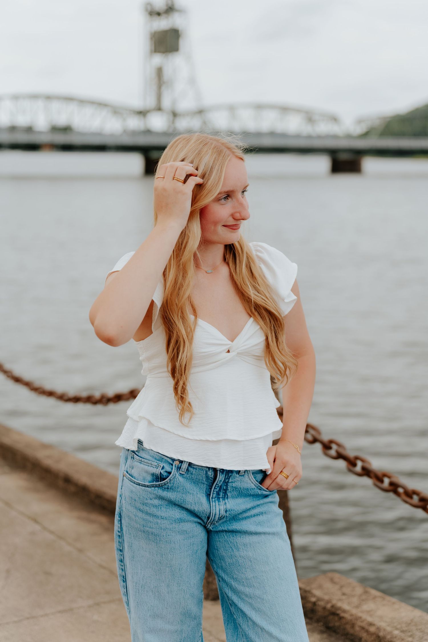 Young blonde woman in white top and jeans poses by a river with a steel bridge in the background.
