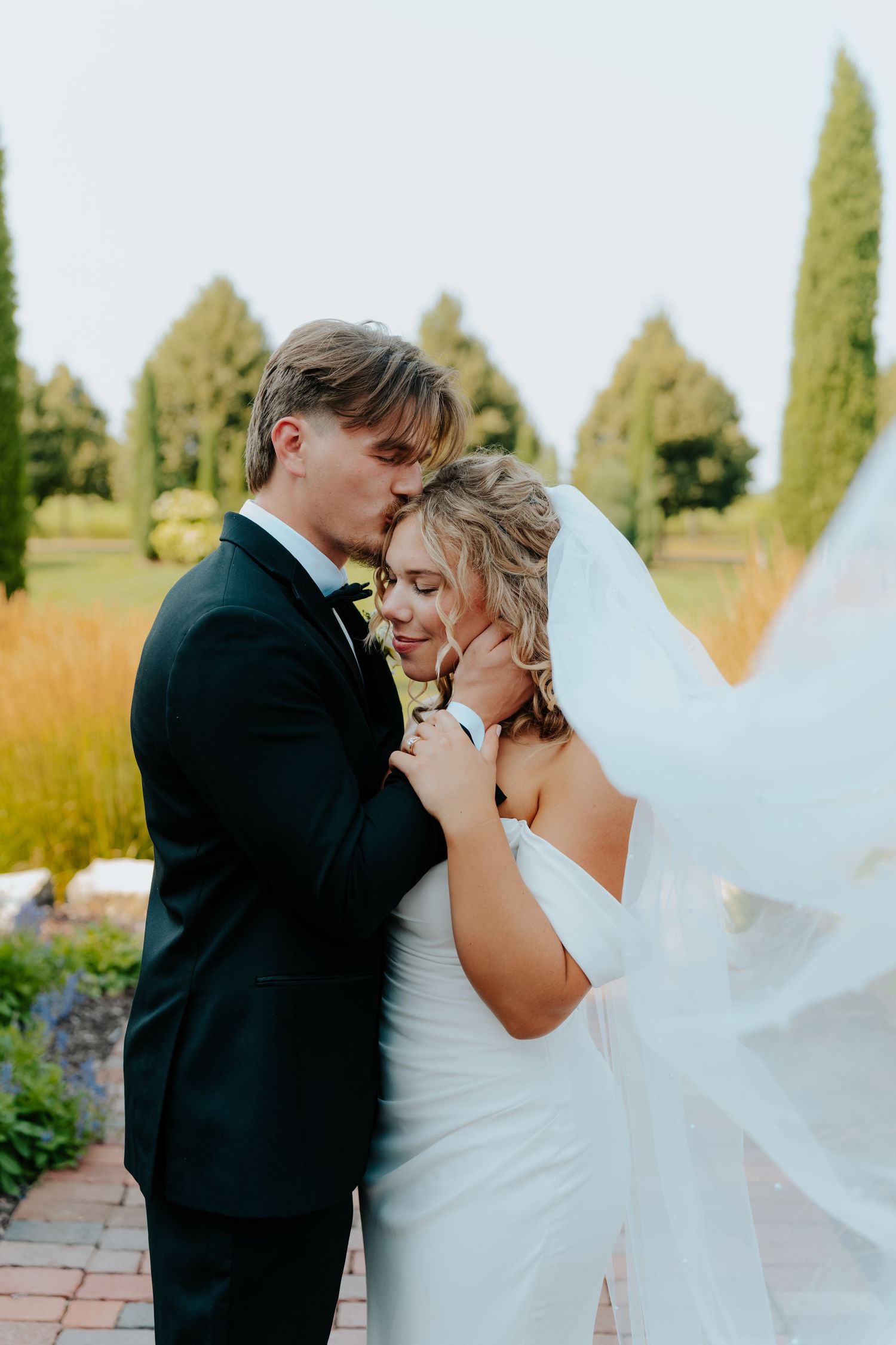 Bride and groom share a romantic moment outdoors, groom kissing bride's forehead as her veil flows in the breeze.