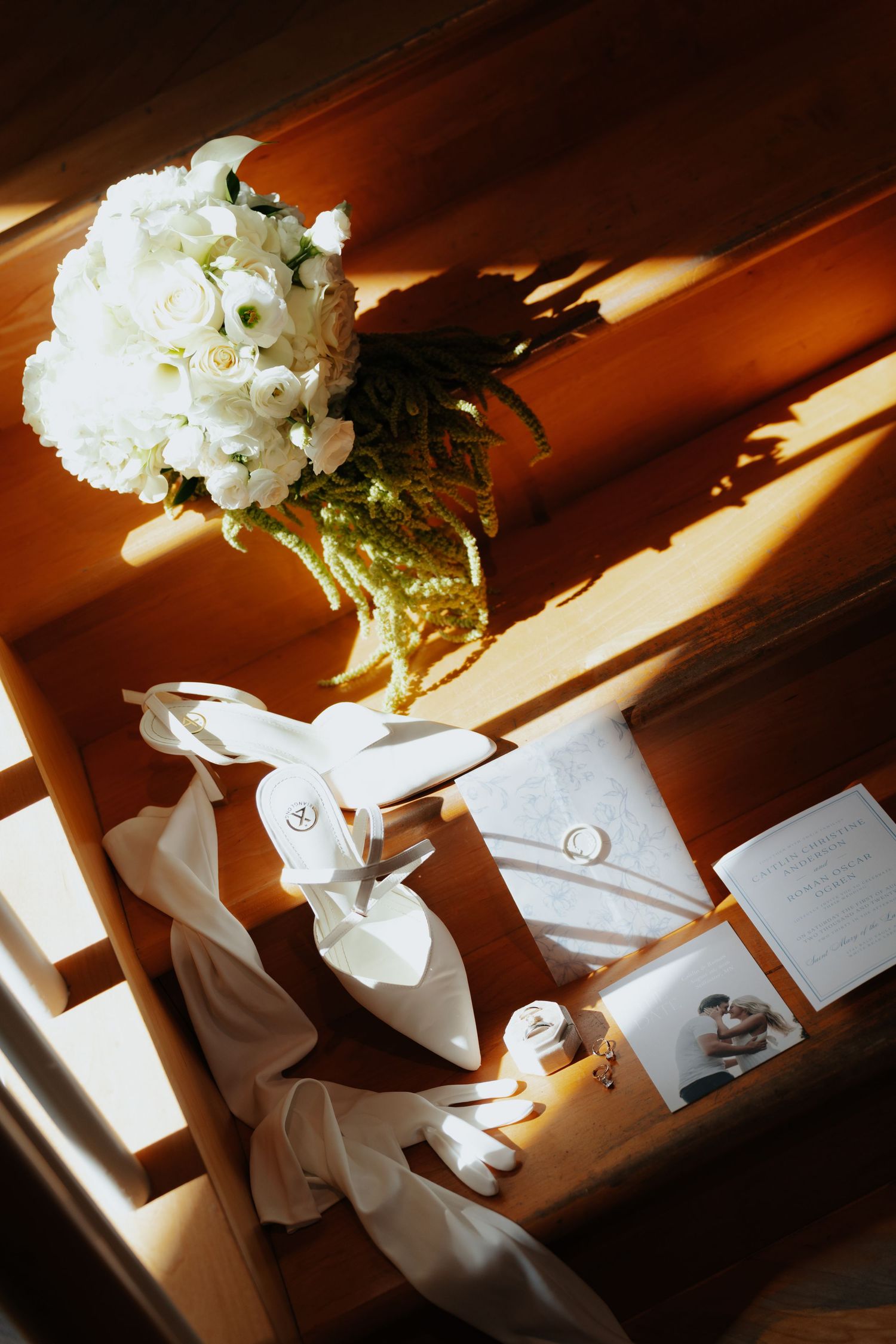 Wedding flat lay featuring white bridal heels, bouquet of white roses, rings, and invitation in warm sunlight.