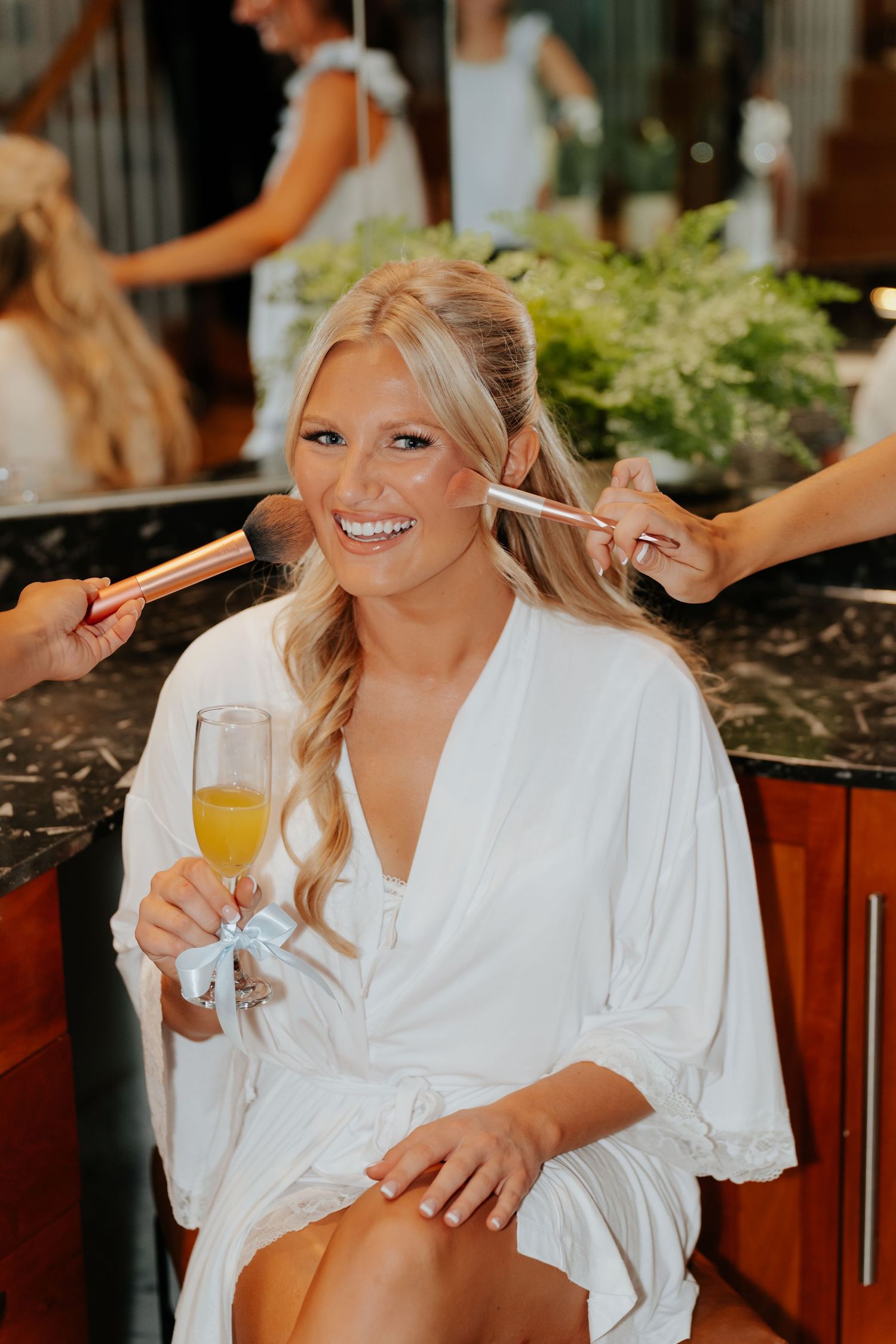 Smiling bride in white robe holds mimosa while getting makeup applied with brushes on wedding day.