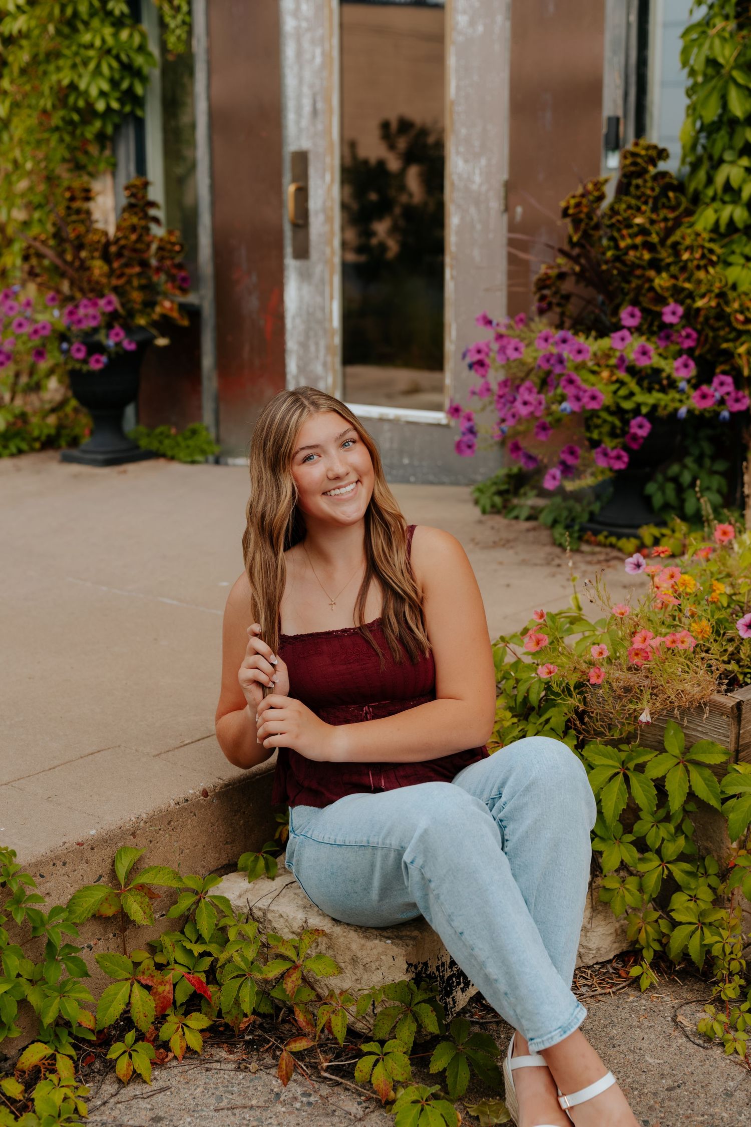 Young woman in burgundy top and jeans sits smiling on steps surrounded by colorful flowers and lush greenery.