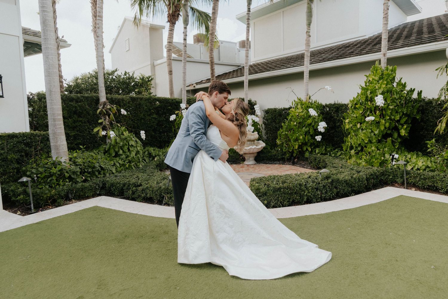 Groom dips bride in white wedding gown outdoors on green lawn surrounded by lush hedges and palm trees.