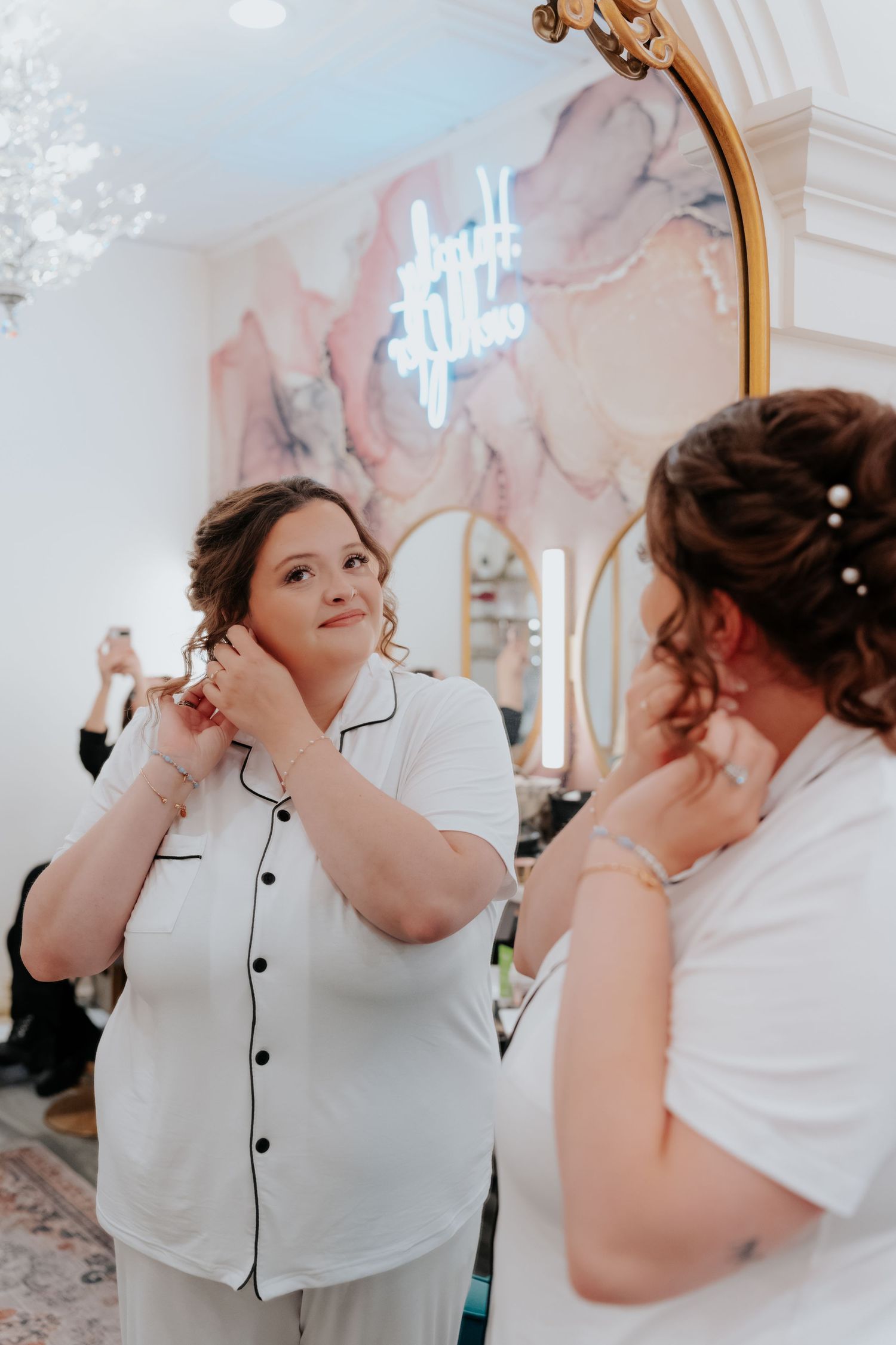 Bride in white pajamas putting on earrings while getting ready in a elegant bridal suite with neon sign.