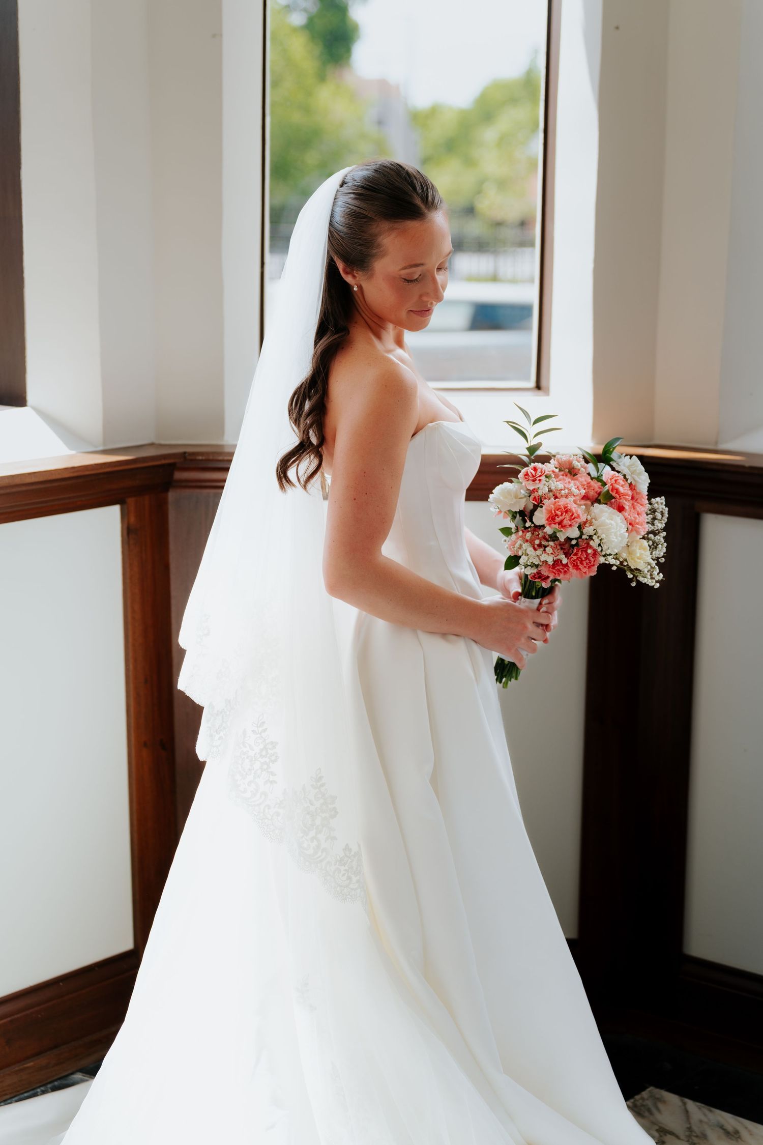 Bride in white strapless gown and veil holds pink bouquet by a bright window on her wedding day.