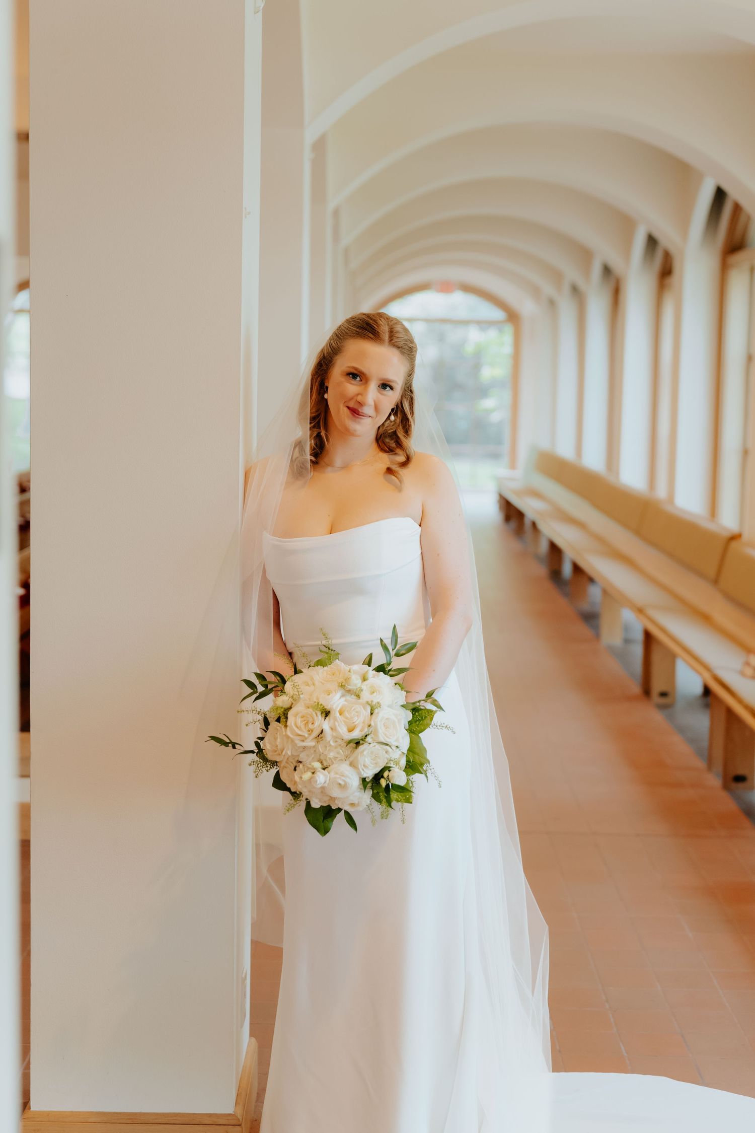 Elegant bride in strapless white gown holding bouquet of white roses in sunlit church corridor.