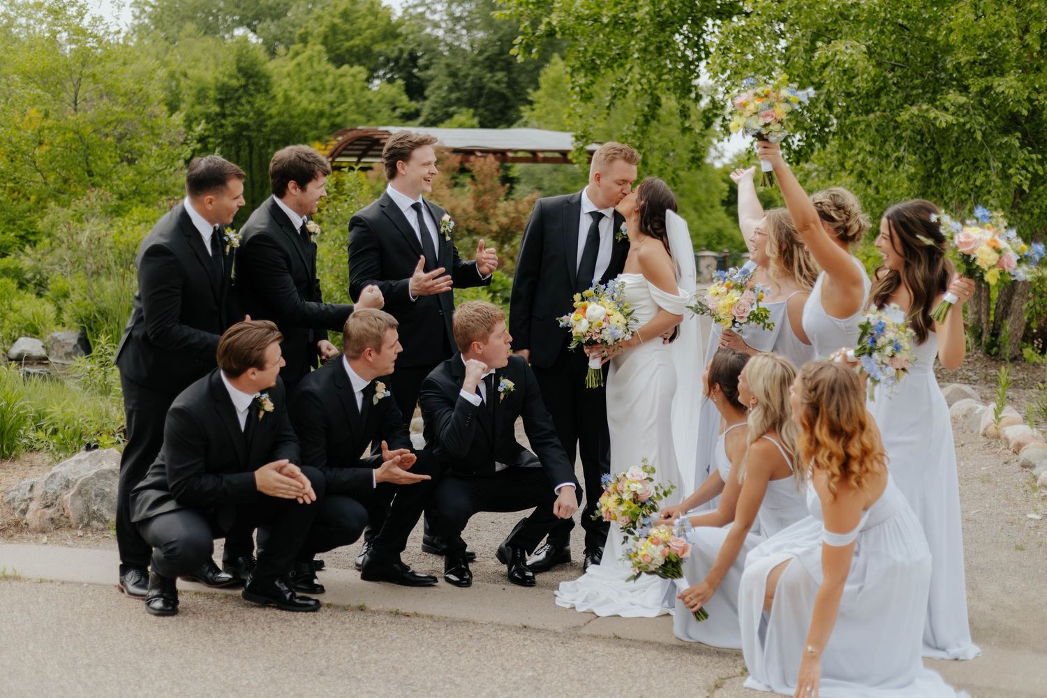 Wedding party in black suits and white dresses celebrating outdoors, with bridesmaids cheering and groomsmen laughing together.