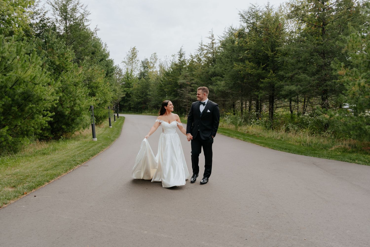 Bride and groom walk hand-in-hand down a tree-lined path, bride in white gown, groom in black tuxedo.