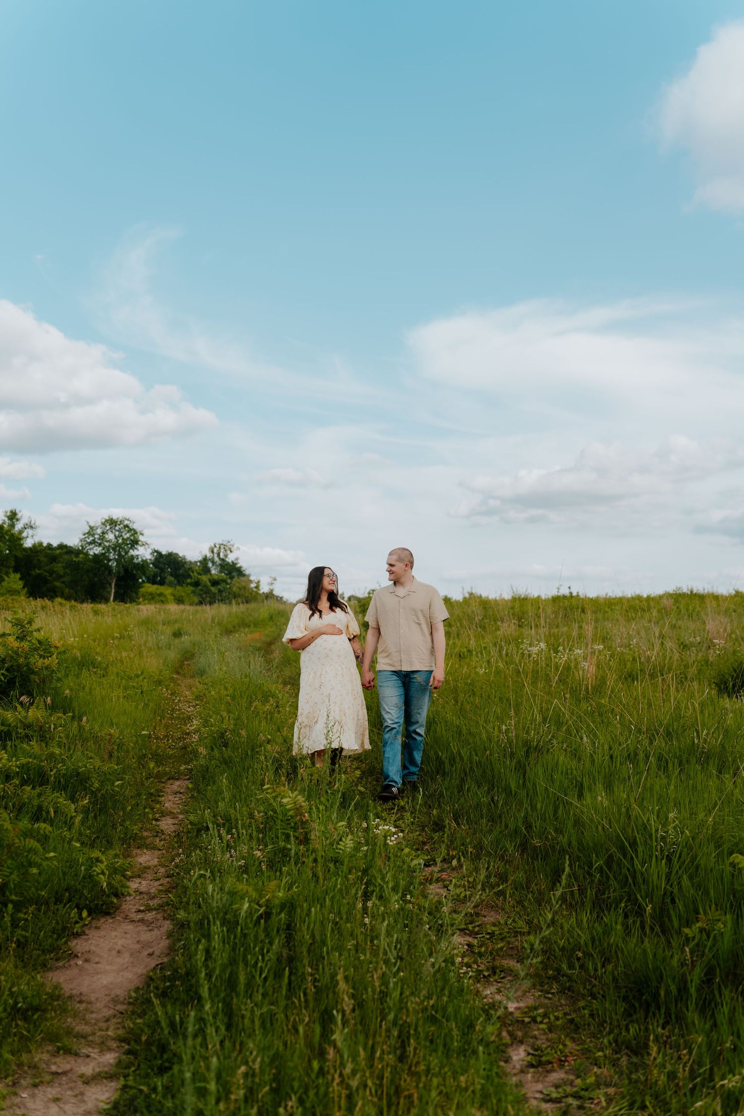 A couple holds hands while walking through a lush green field under a blue sky.
