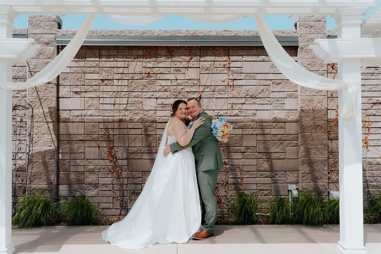 Bride in white gown and groom in sage suit embrace under white pergola against stone wall backdrop.