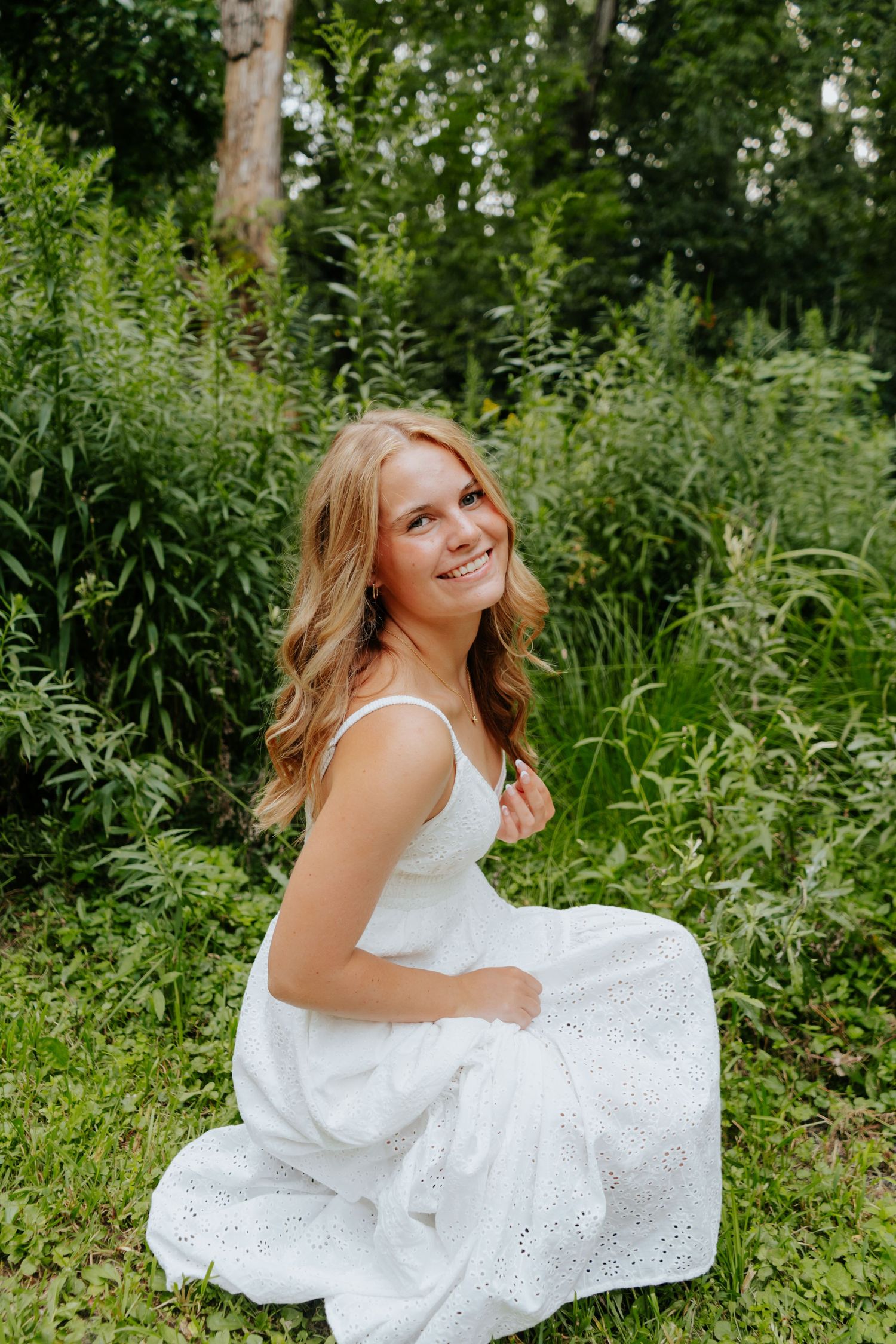 Smiling young woman with blonde wavy hair wearing a white sundress sitting outdoors among lush green plants.