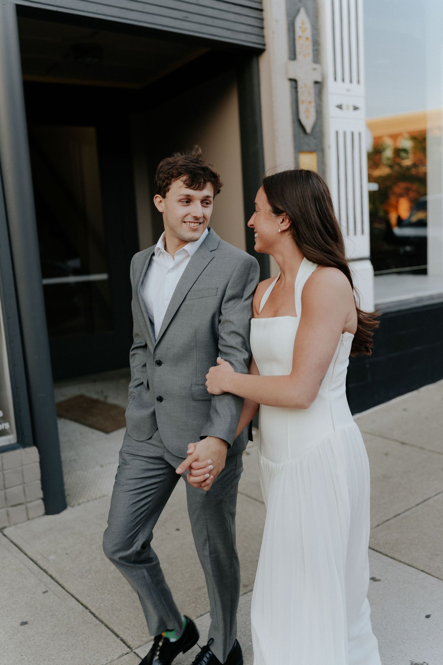A couple in wedding attire holds hands and smiles while walking on a city sidewalk, groom in gray suit, bride in white dress.