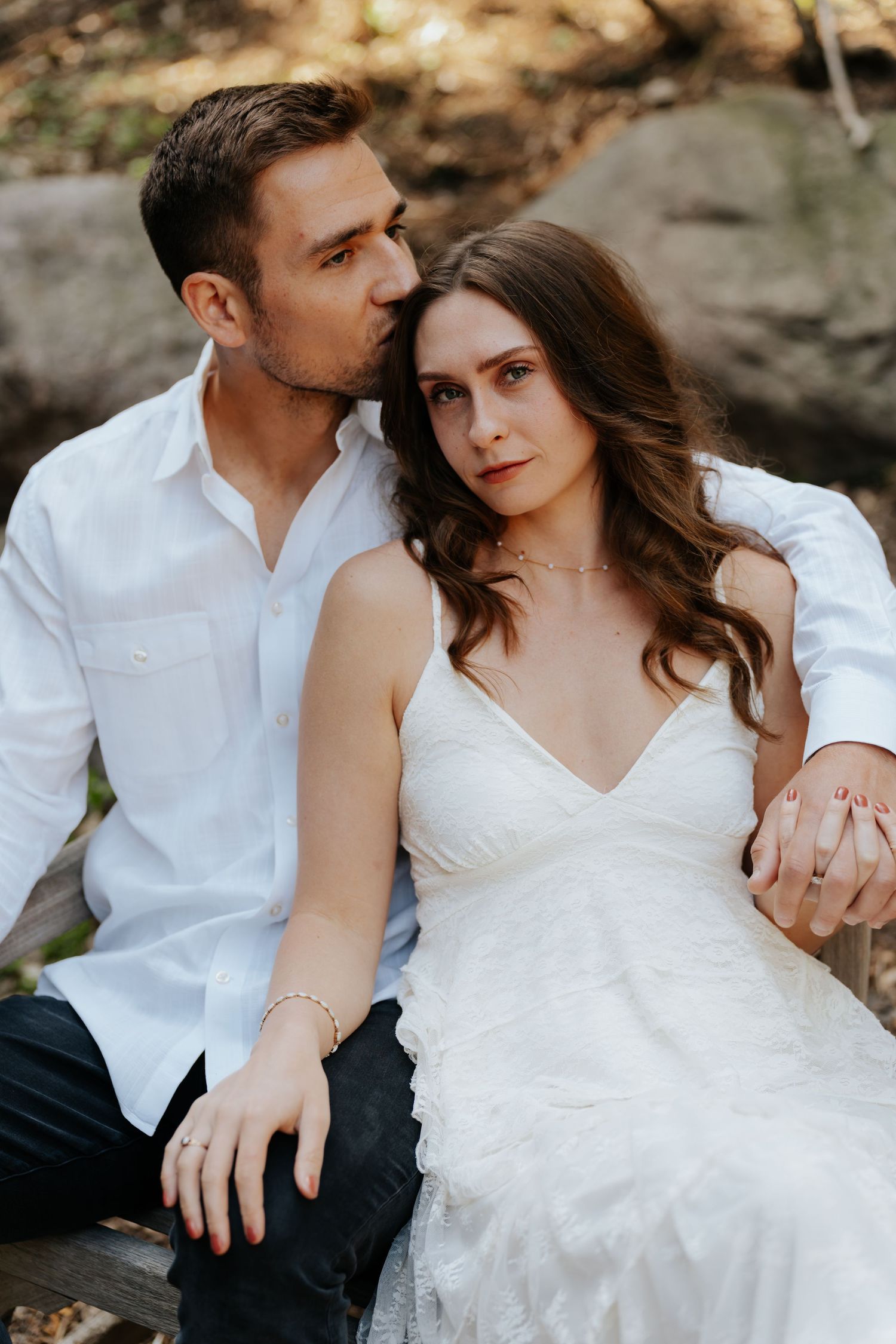 A couple in white attire sits closely outdoors, the man kissing the woman's head as she gazes forward serenely.