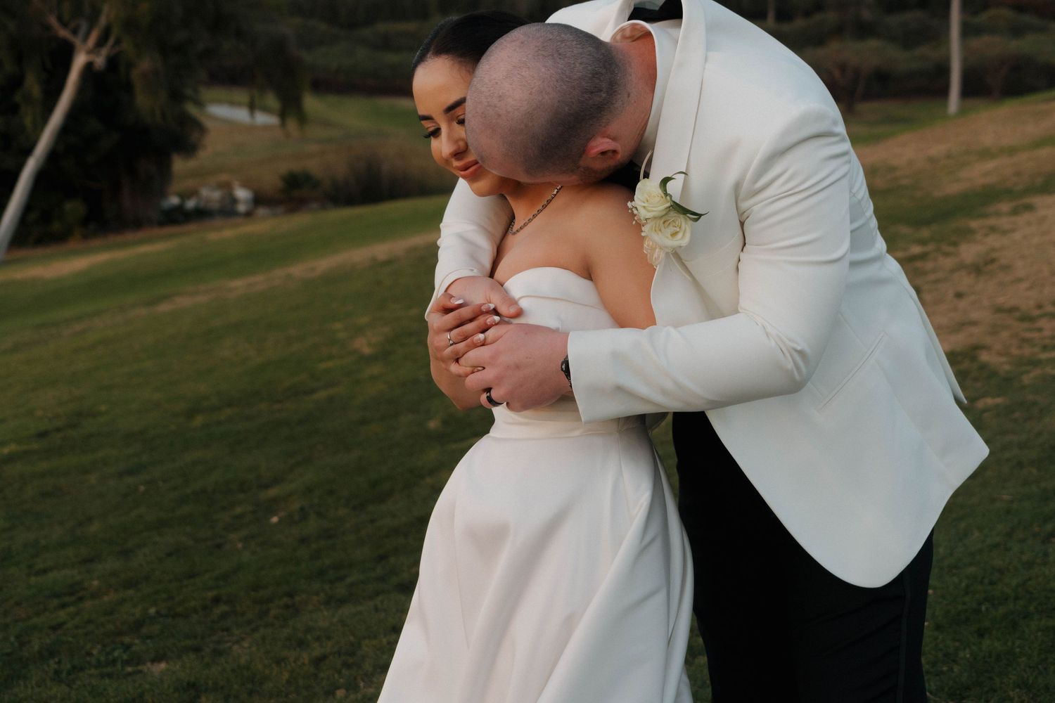 A couple in wedding attire embrace during sunset in a grassy outdoor setting.