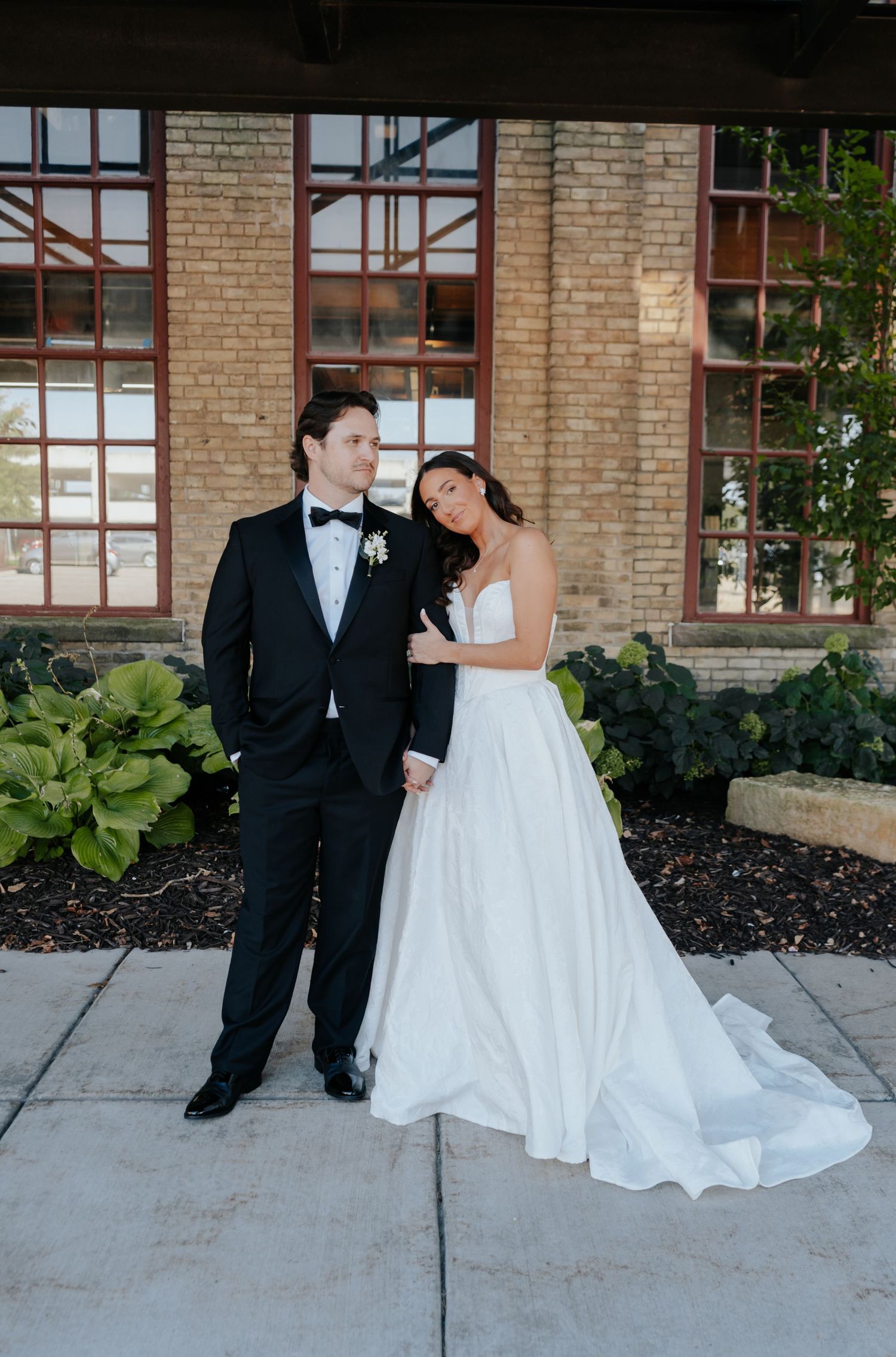 Wedding couple poses against a rustic brick building with large windows.