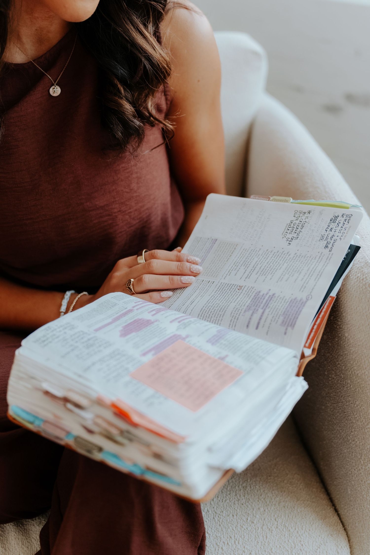 Woman in burgundy dress reading a heavily annotated Bible with colorful highlights while sitting on a beige sofa.