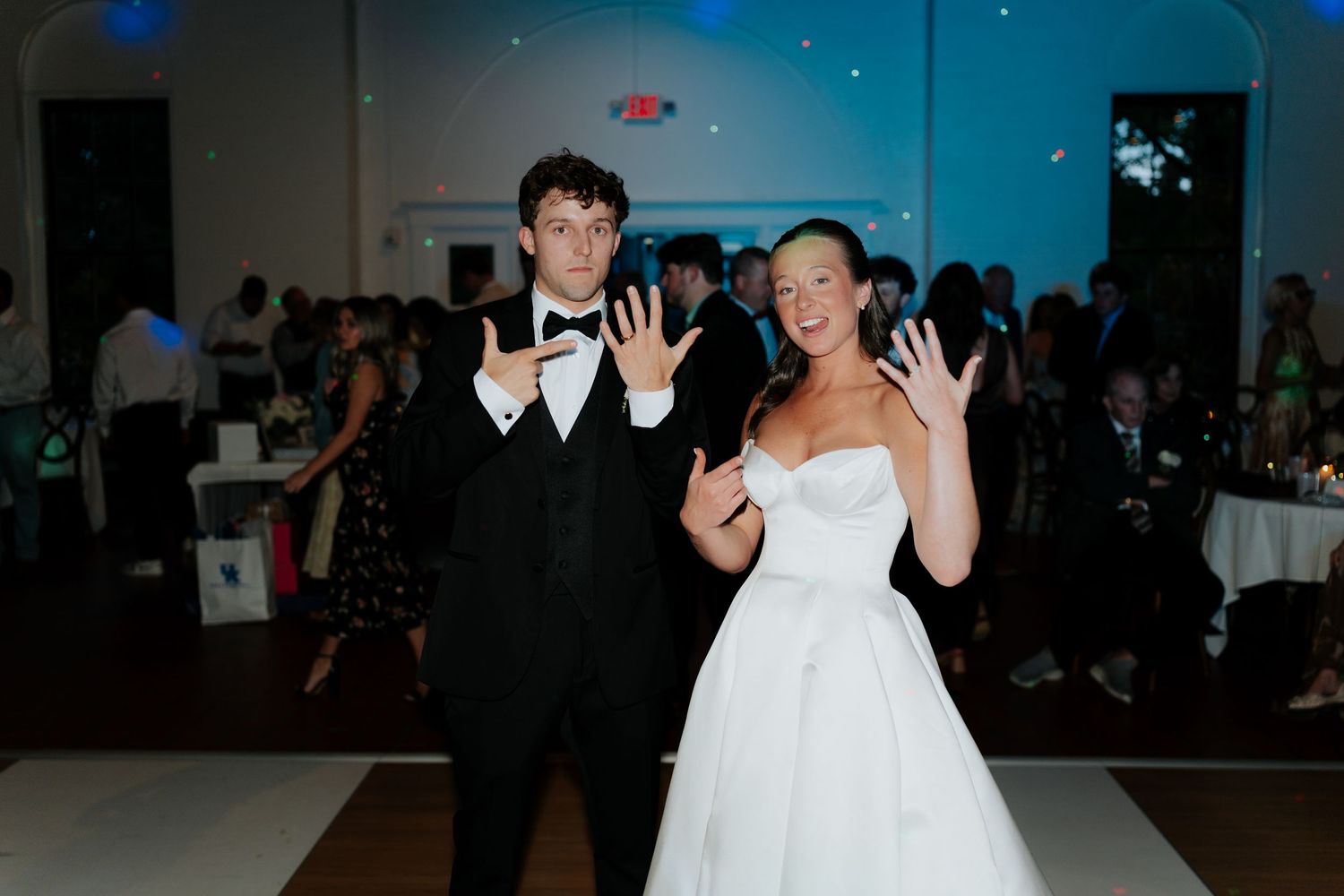 Bride and groom showing off wedding rings on dance floor at elegant wedding reception with blue lighting.