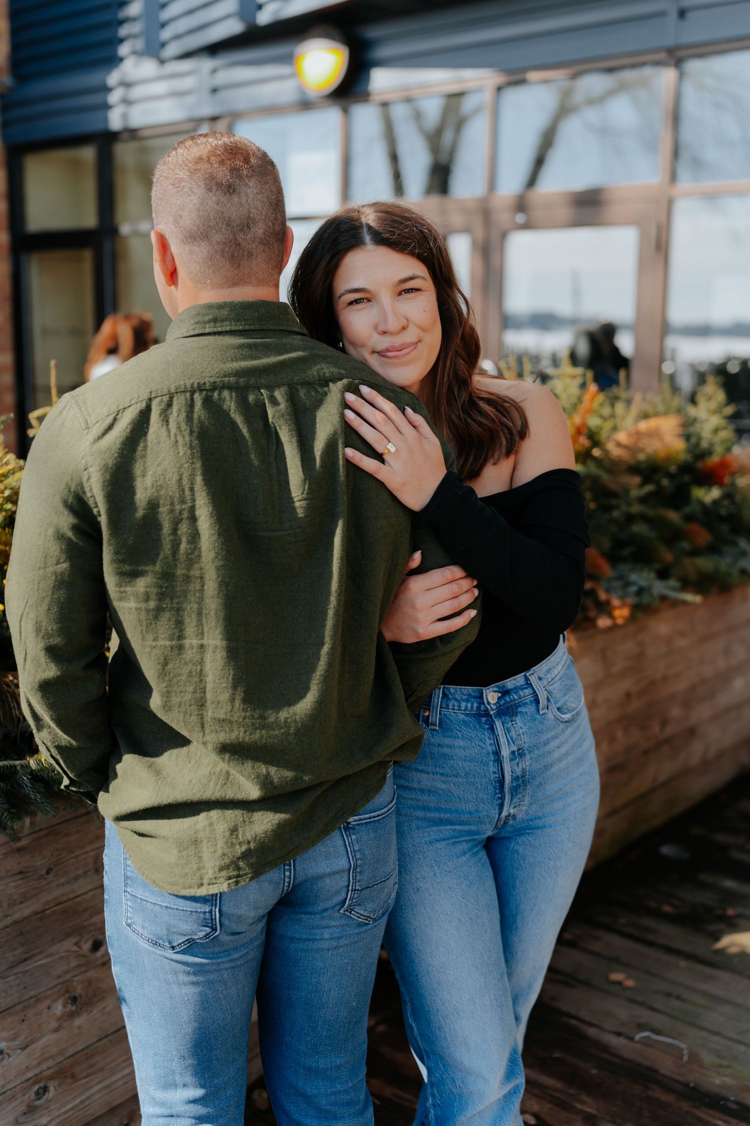 A couple embraces outdoors, woman in black off-shoulder top and jeans smiling, man in green shirt, fall flowers behind them.