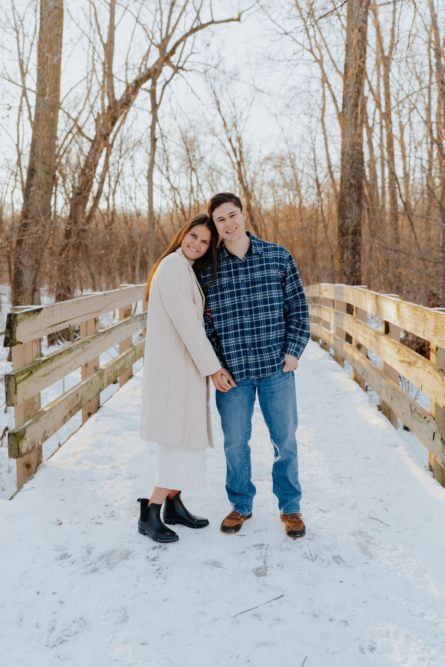 Couple posing on a snowy wooden bridge in winter, woman in cream coat and man in plaid shirt holding hands.