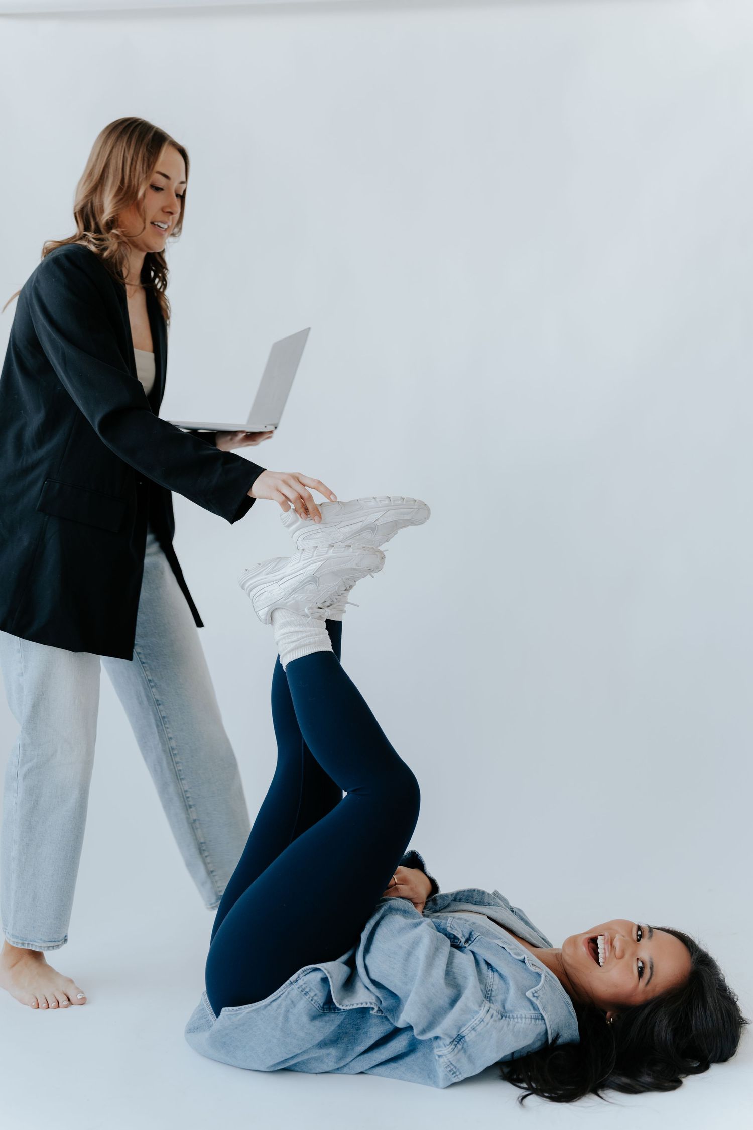 Two women laughing playfully in a studio, one holding a laptop while the other poses on the floor.