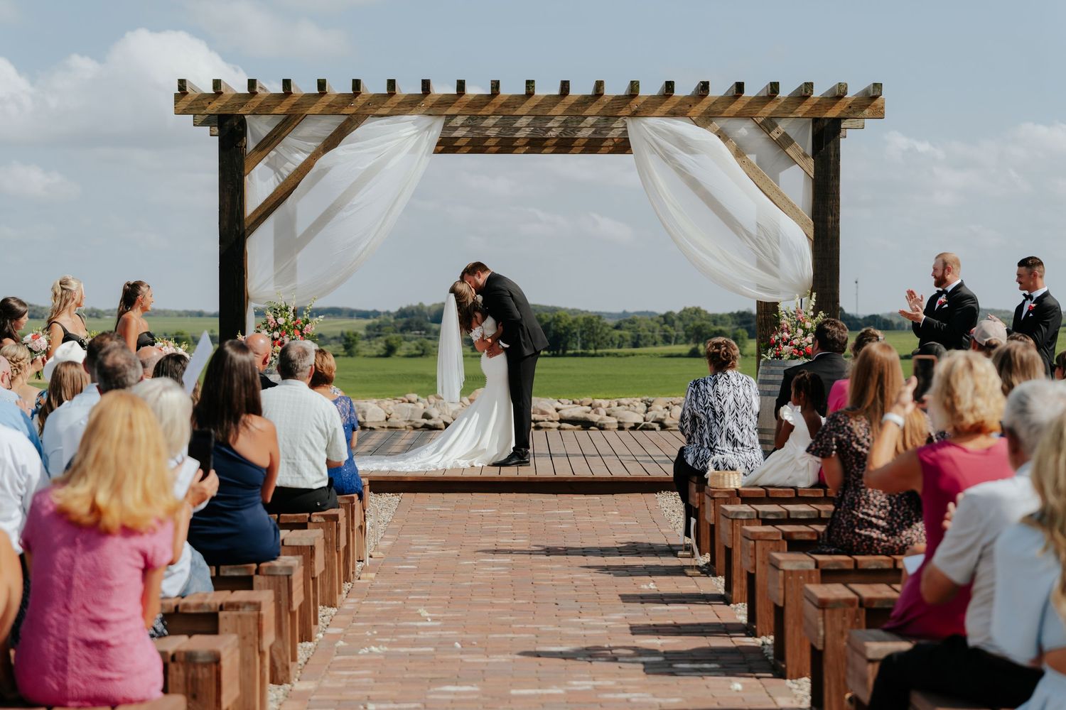 Outdoor wedding ceremony under rustic wooden pergola with white draping, couple kissing at altar, guests seated on wooden pews.