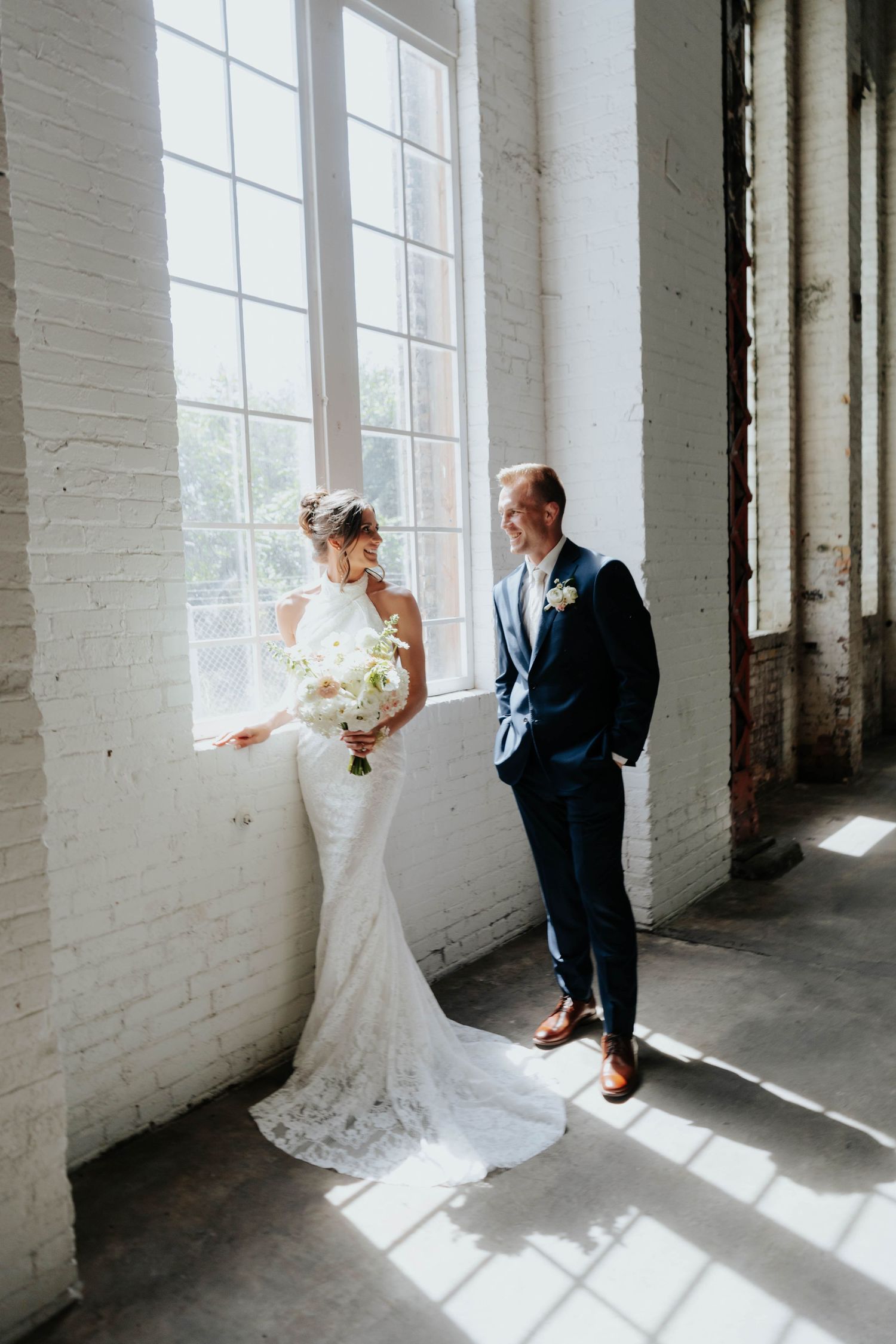 Bride in lace gown holding white bouquet stands with groom in navy suit by large window in rustic venue.