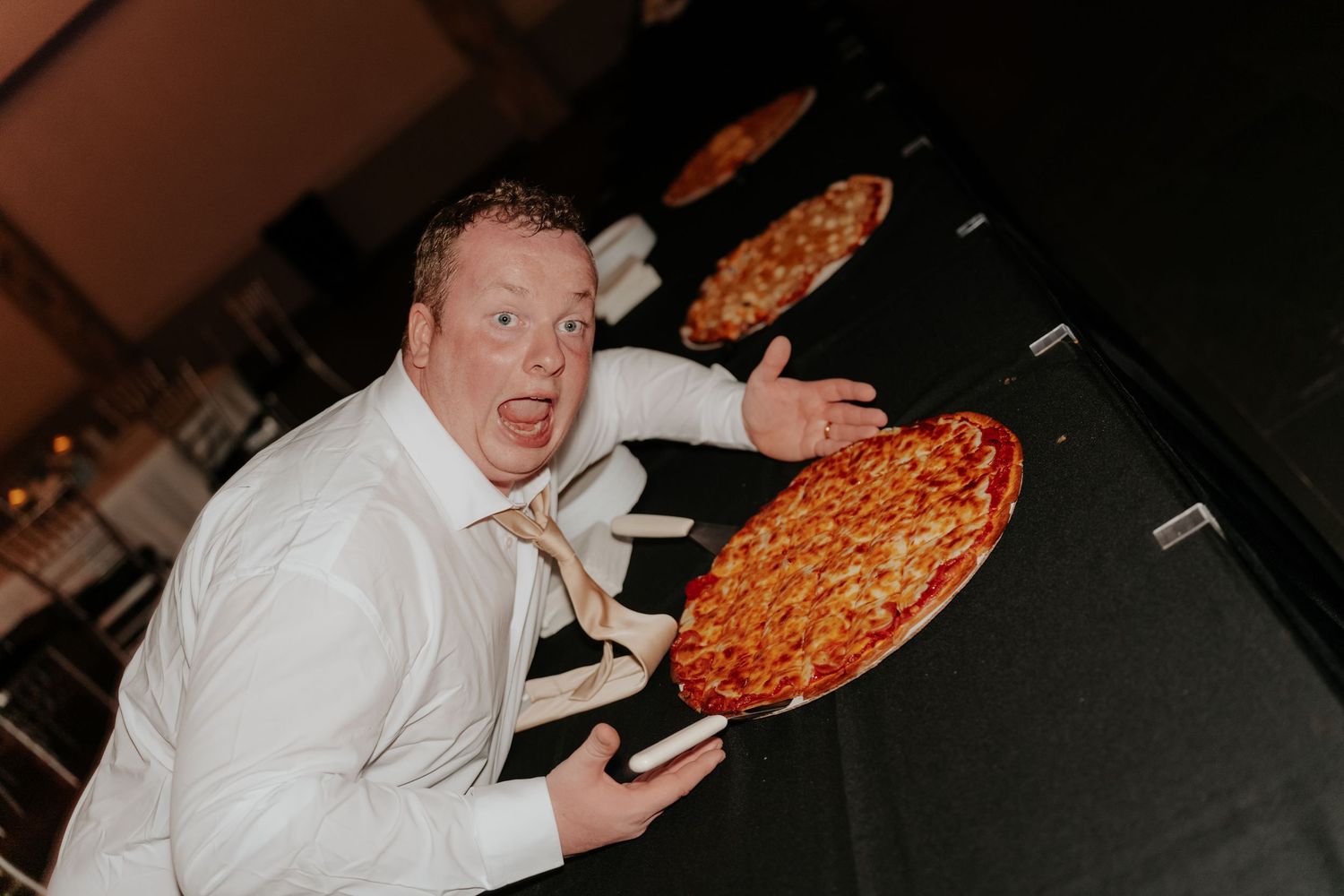Excited man in white shirt tossing a pepperoni pizza in the air at a wedding reception.
