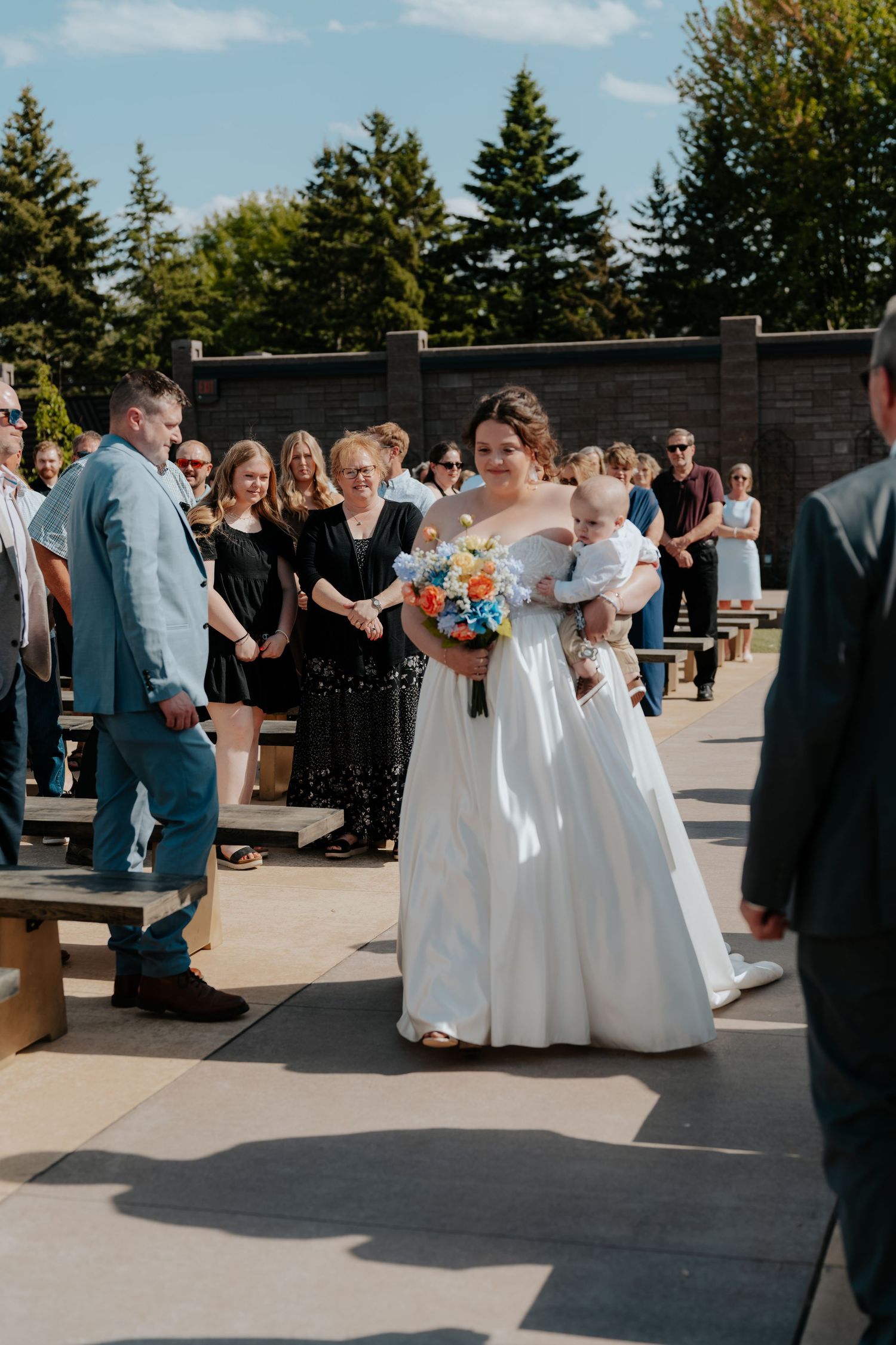 Bride in elegant white off-shoulder gown walks down outdoor aisle holding colorful bouquet and baby at sunny wedding.