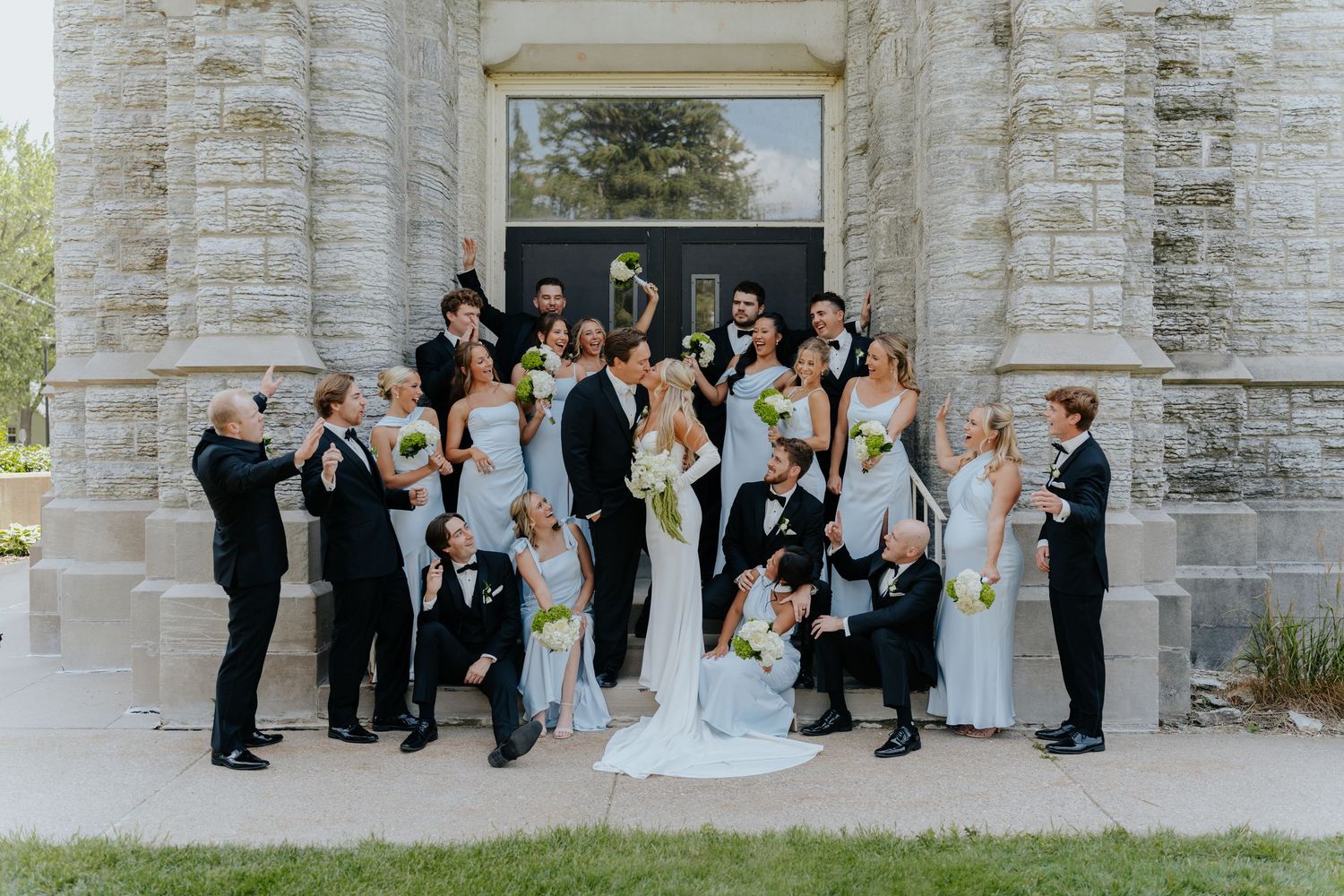 Wedding party poses joyfully outside a stone church, with bridesmaids in light blue gowns and groomsmen in black tuxedos.