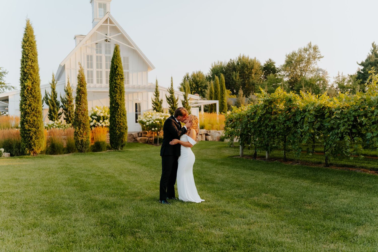 Couple kisses in front of white chapel surrounded by lush gardens.