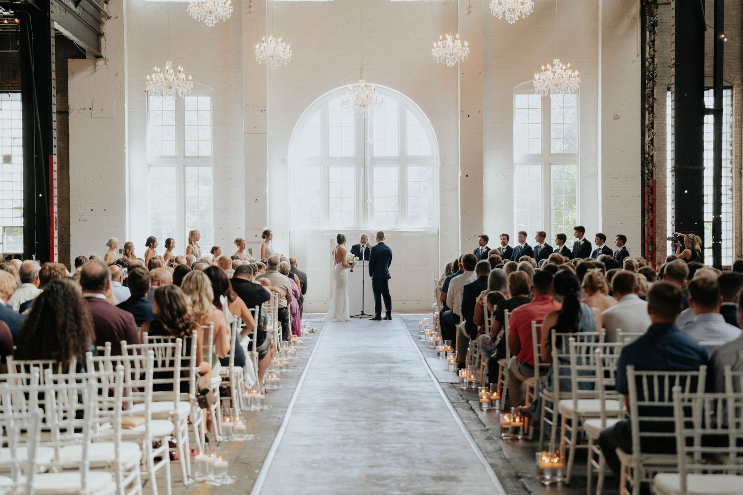 Elegant wedding ceremony in bright ballroom with chandelier, white chairs, and guests seated along aisle.