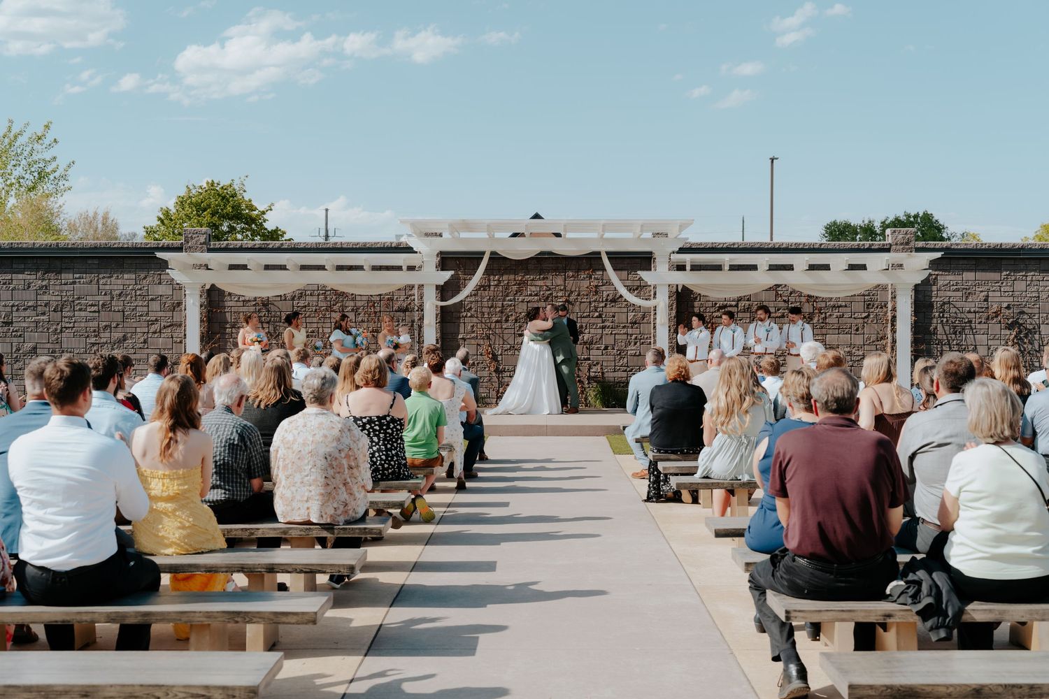 Guests seated on wooden benches watch a wedding ceremony at an outdoor venue with a white pergola altar.