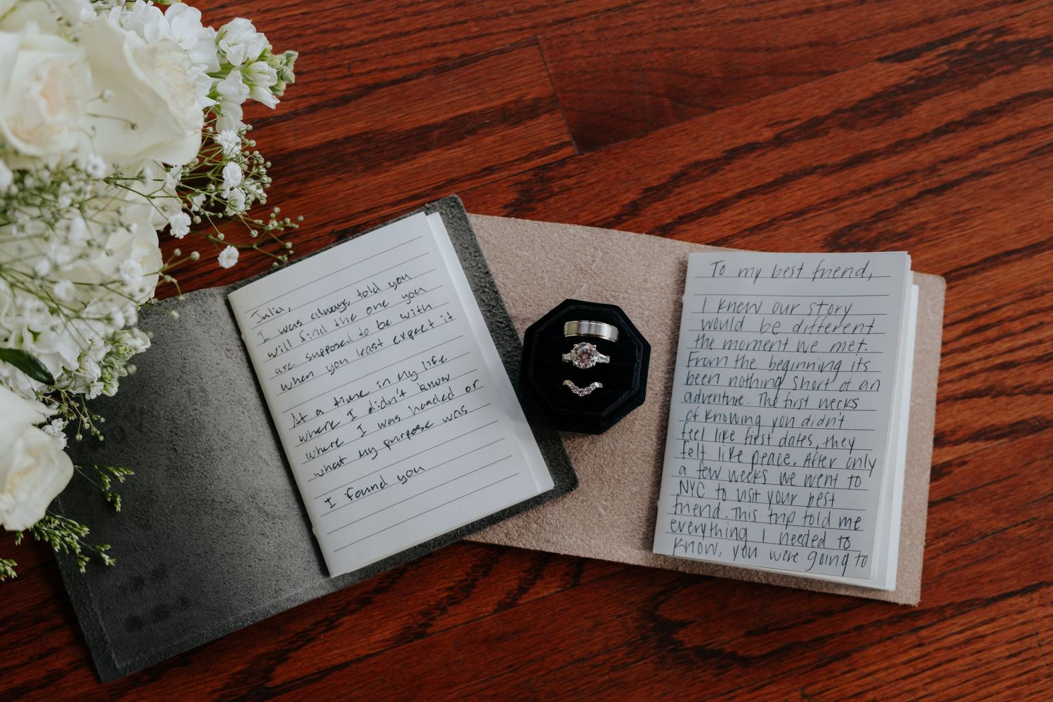 Wedding vow books open with handwritten notes, diamond engagement ring in black velvet box on wooden table with white flowers.