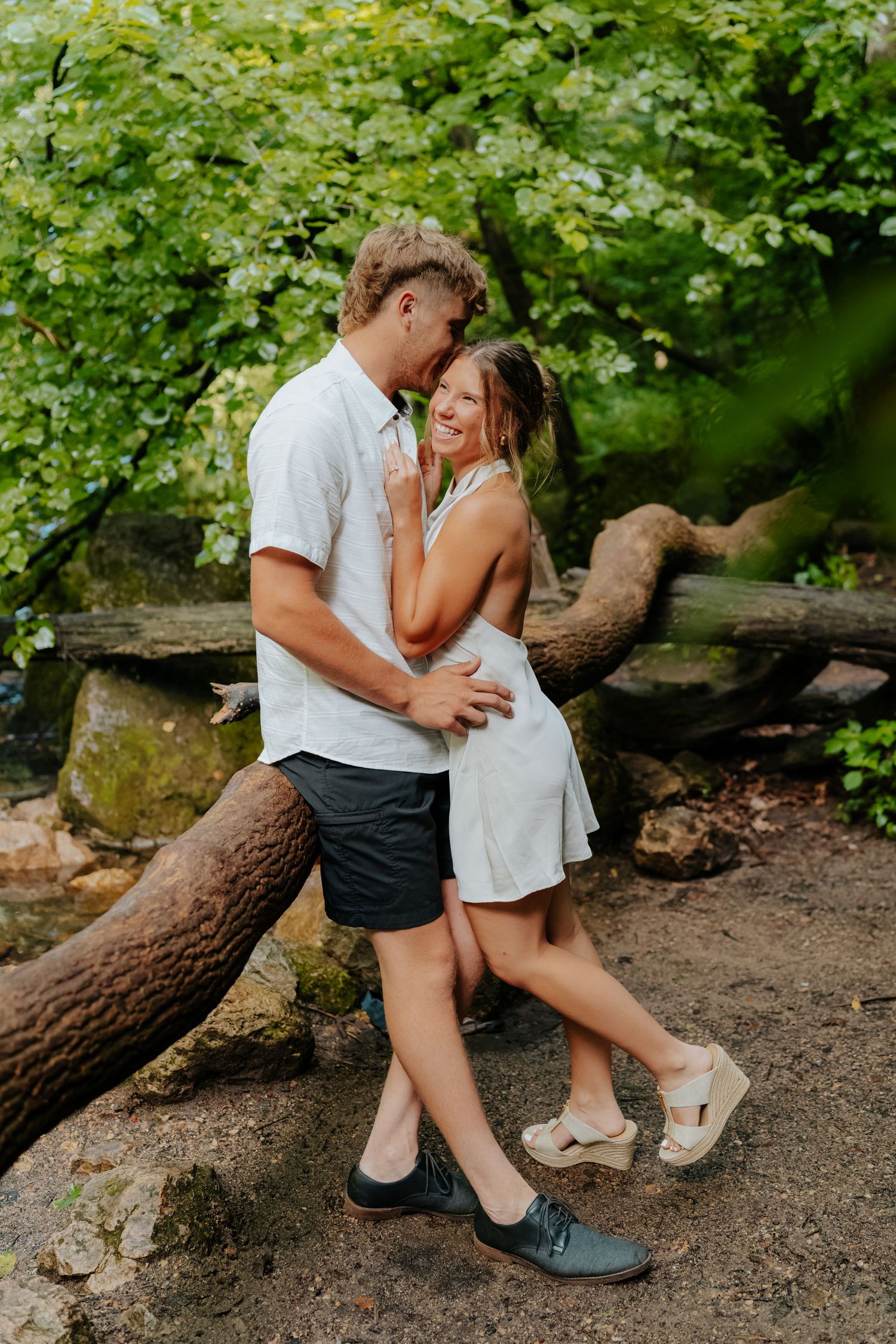 A couple embraces and smiles near a fallen tree surrounded by lush green foliage outdoors.