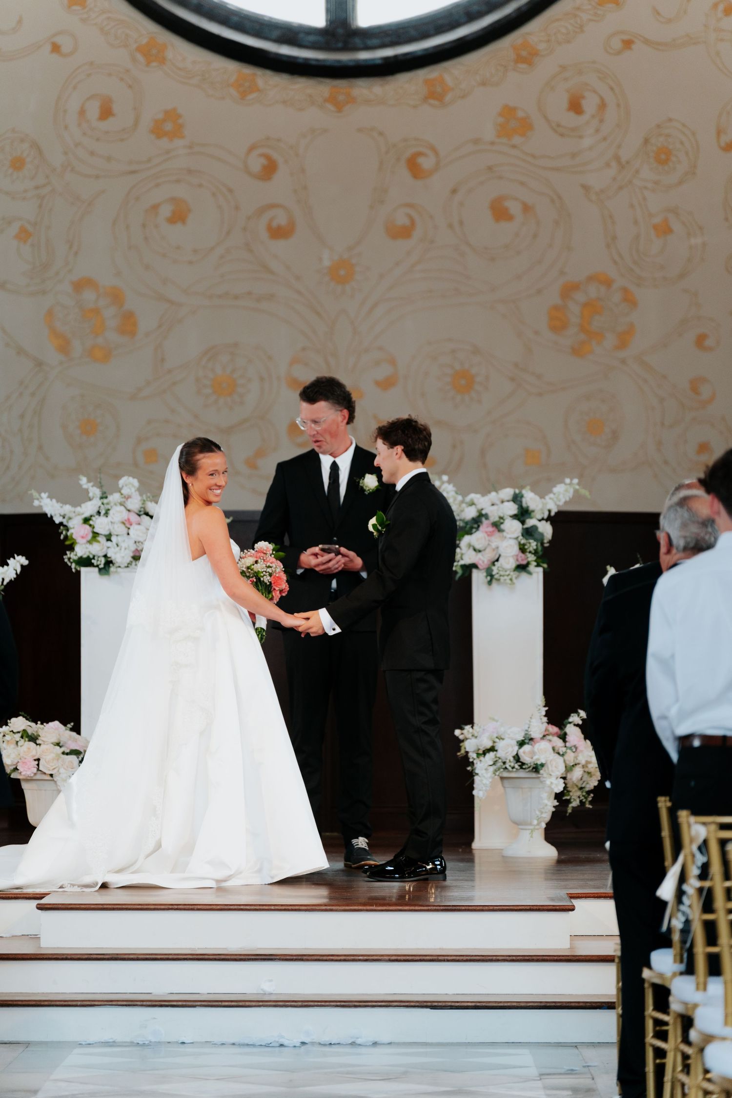 Bride and groom exchange vows at elegant wedding ceremony with white floral arrangements and ornate gold wall decor.