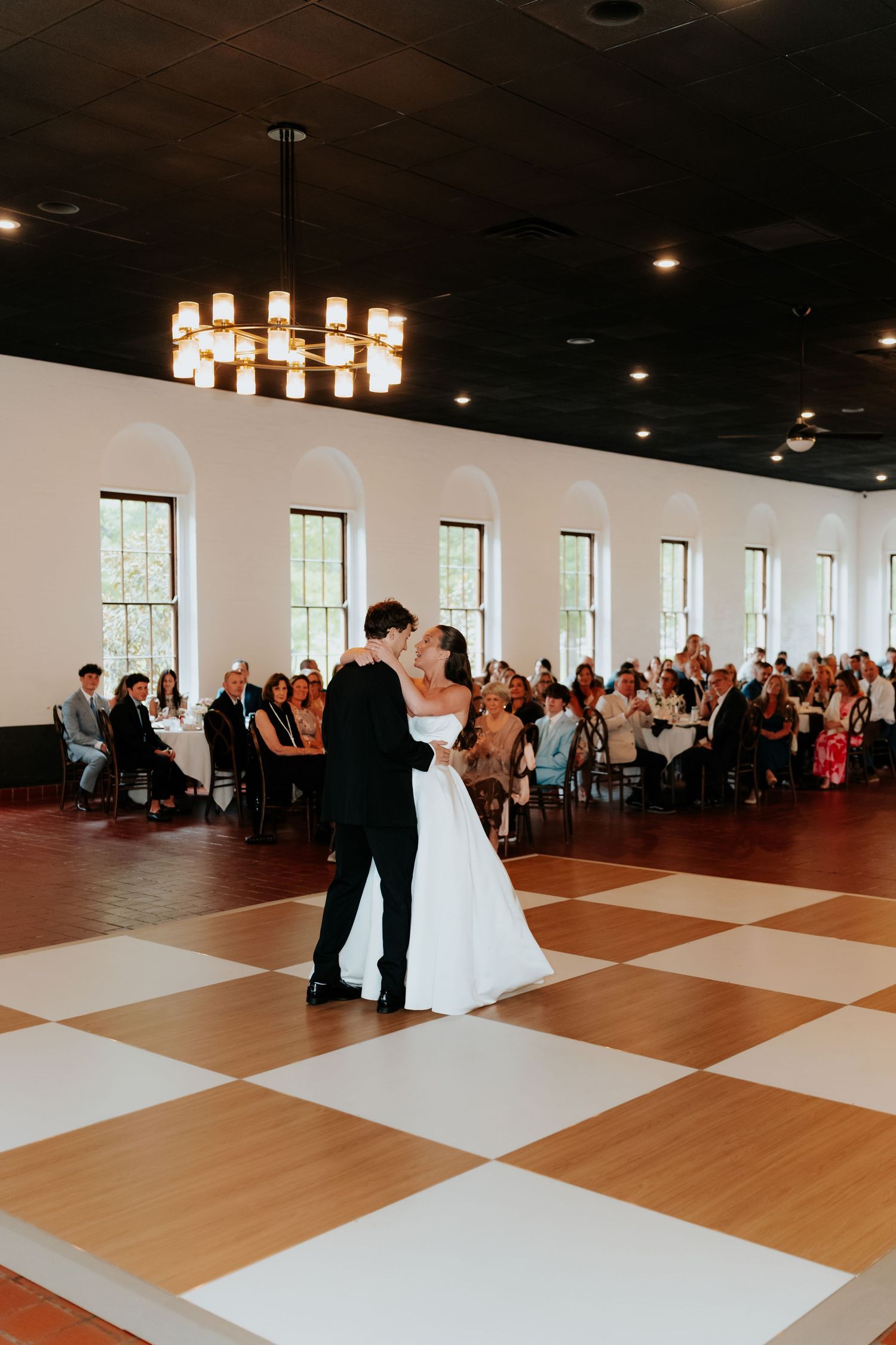 Bride and groom share first dance on checkered floor in elegant ballroom with guests seated around them.