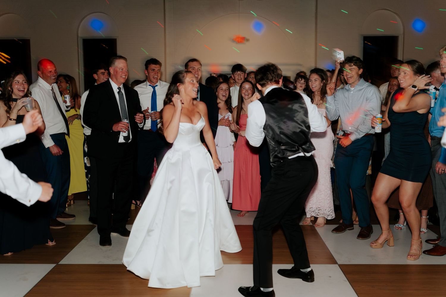Bride and groom share a lively dance with guests at their wedding reception on a checkered dance floor.