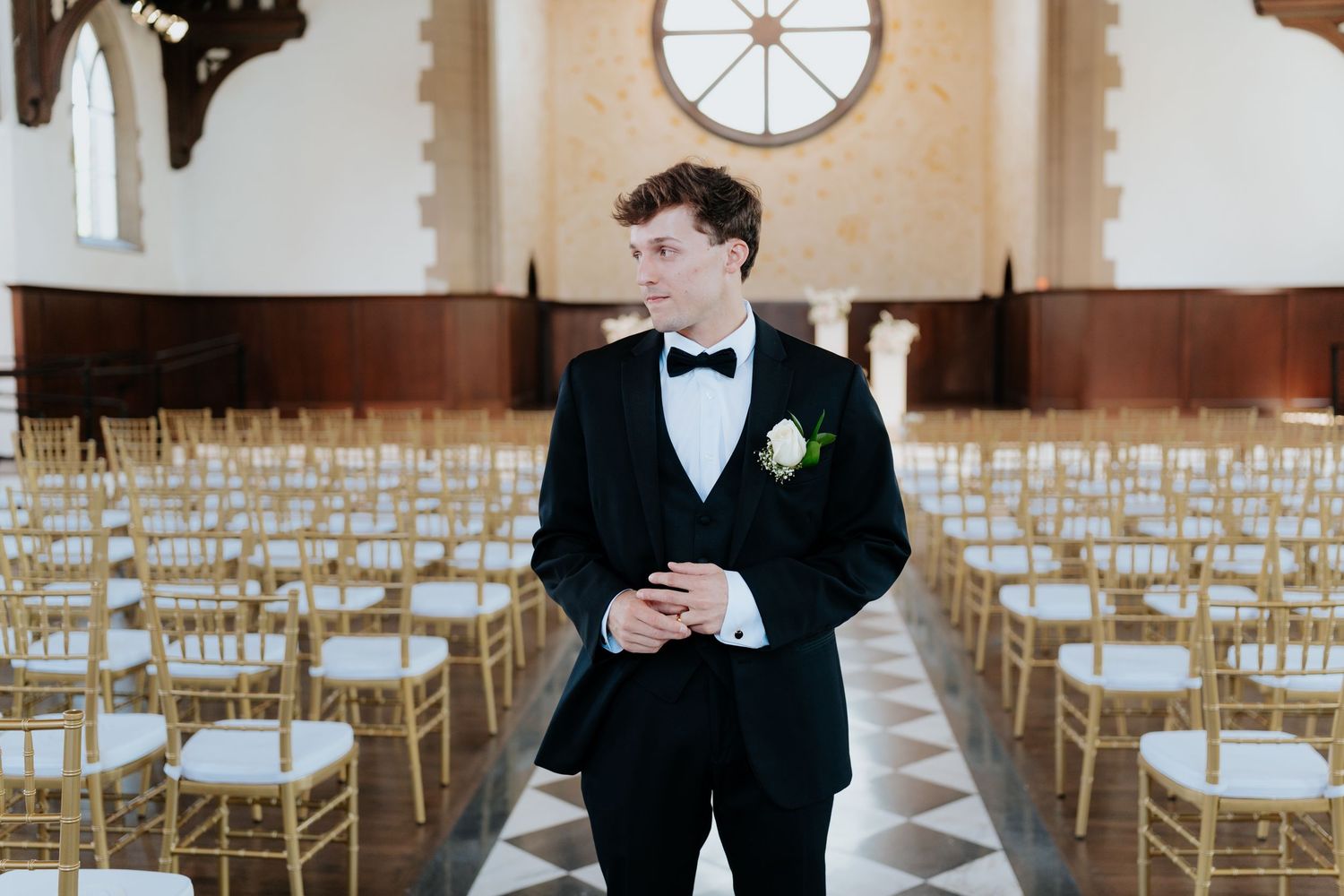 Groom in black tuxedo with boutonniere stands in elegant wedding venue aisle surrounded by gold chiavari chairs.