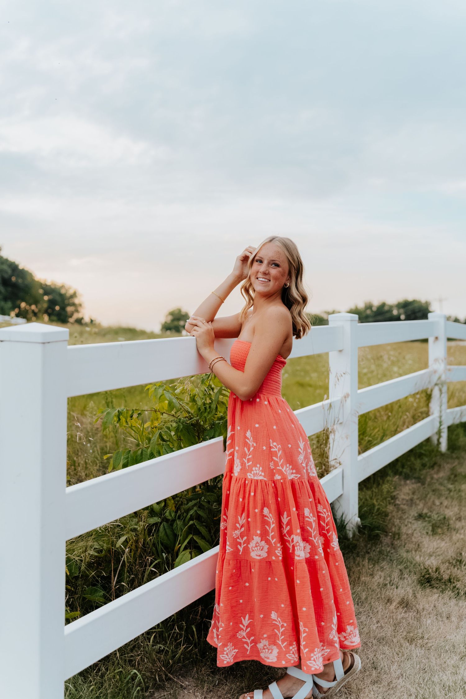 Young woman in coral floral maxi dress poses by white fence in green field at sunset.