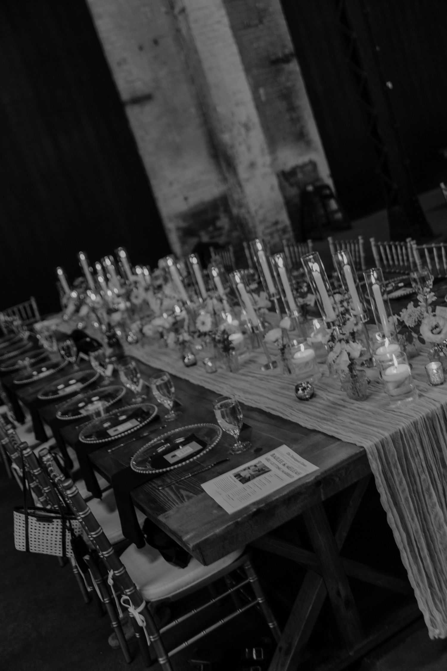 Black and white photo of an elegantly set banquet table with candles, glassware, and place settings in a rustic venue.