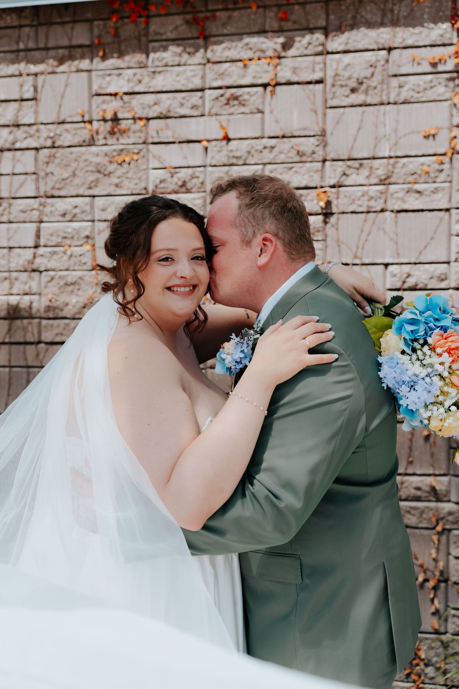 A bride in a white gown and veil smiles as her groom in a sage suit kisses her cheek, holding blue hydrangeas.