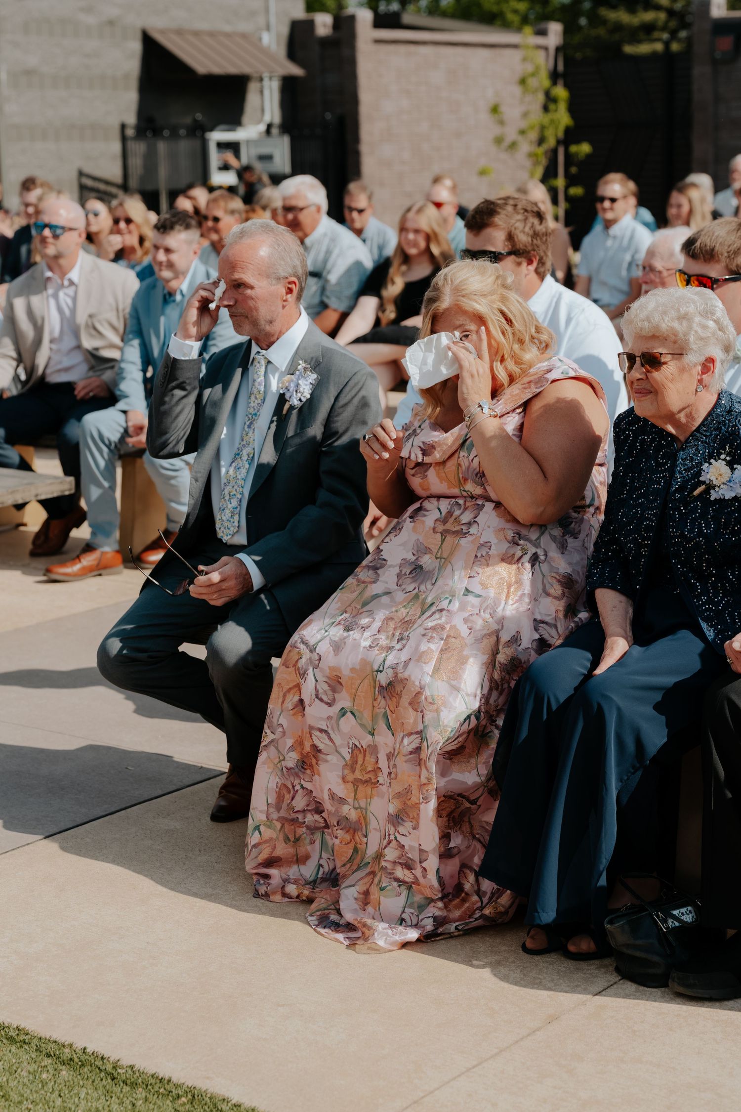 Emotional wedding guests in floral and formal attire wiping tears during outdoor ceremony on sunny day.
