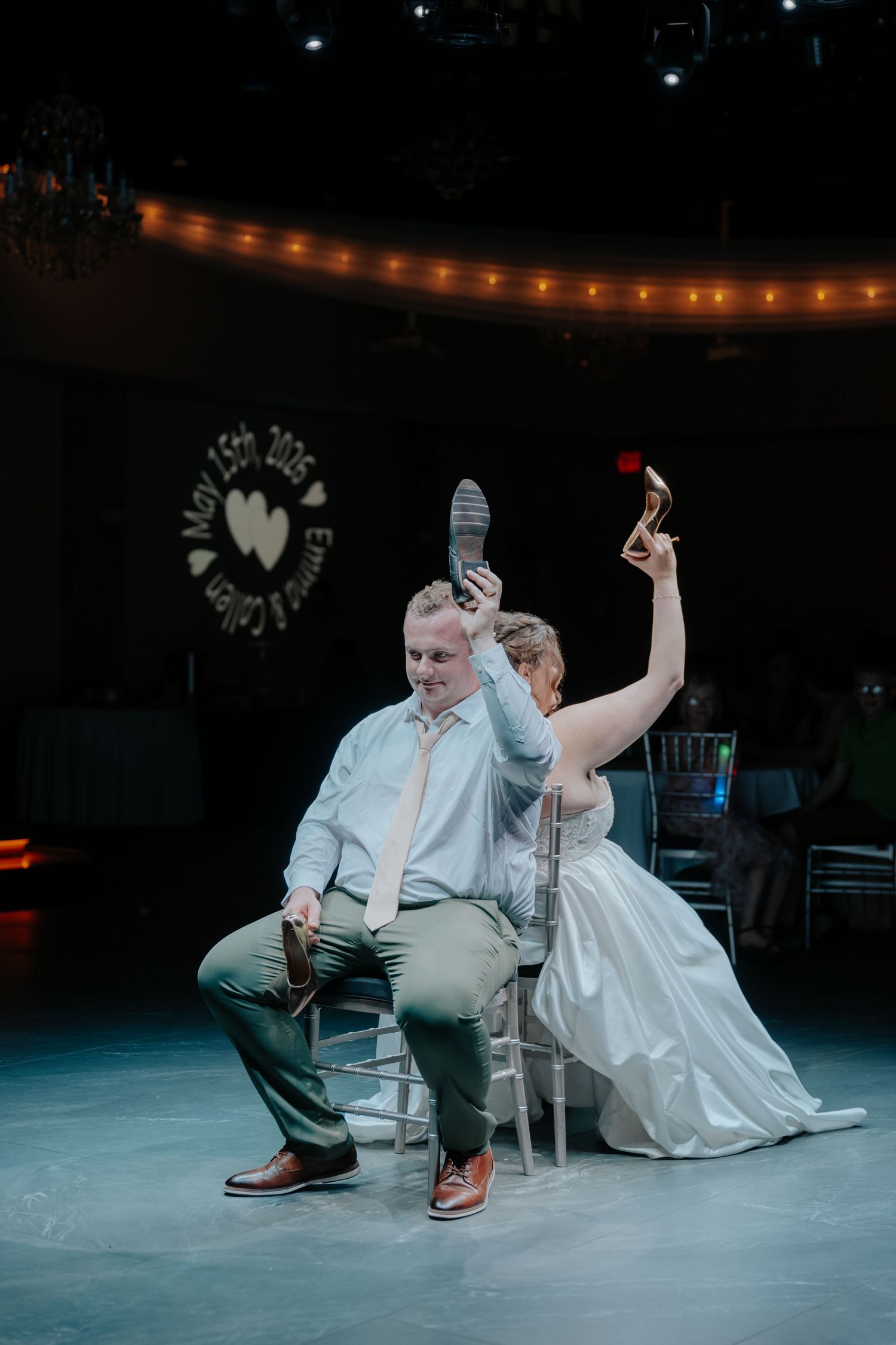 Bride and groom play the shoe game at their wedding reception, holding shoes aloft on a dark stage with string lights.