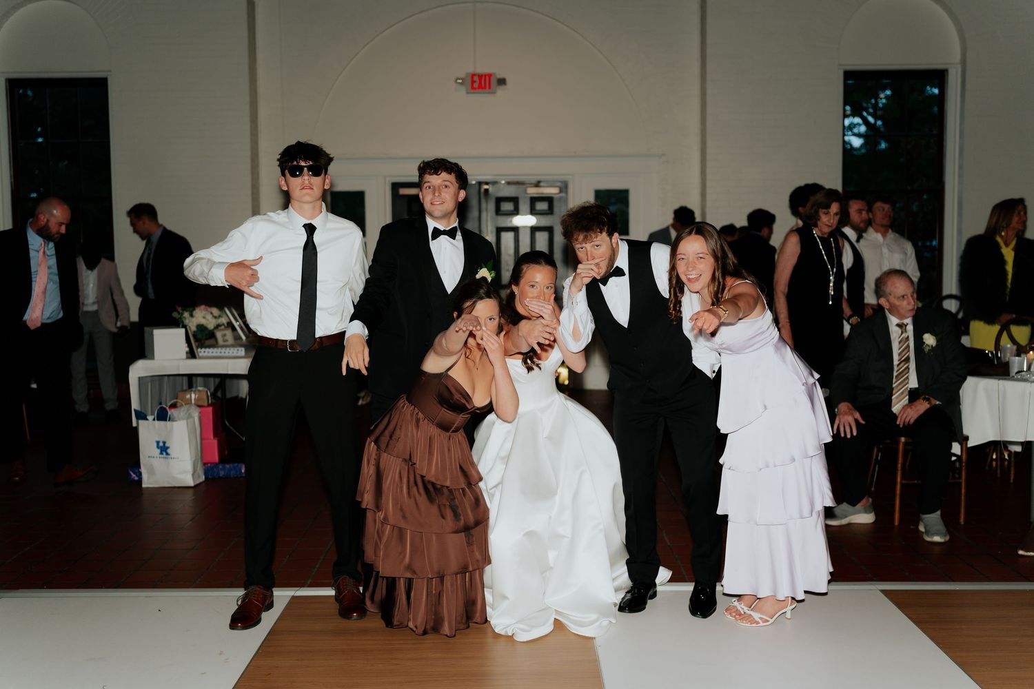 Wedding party posing playfully on dance floor, bride in white gown surrounded by guests in formal attire at elegant reception.