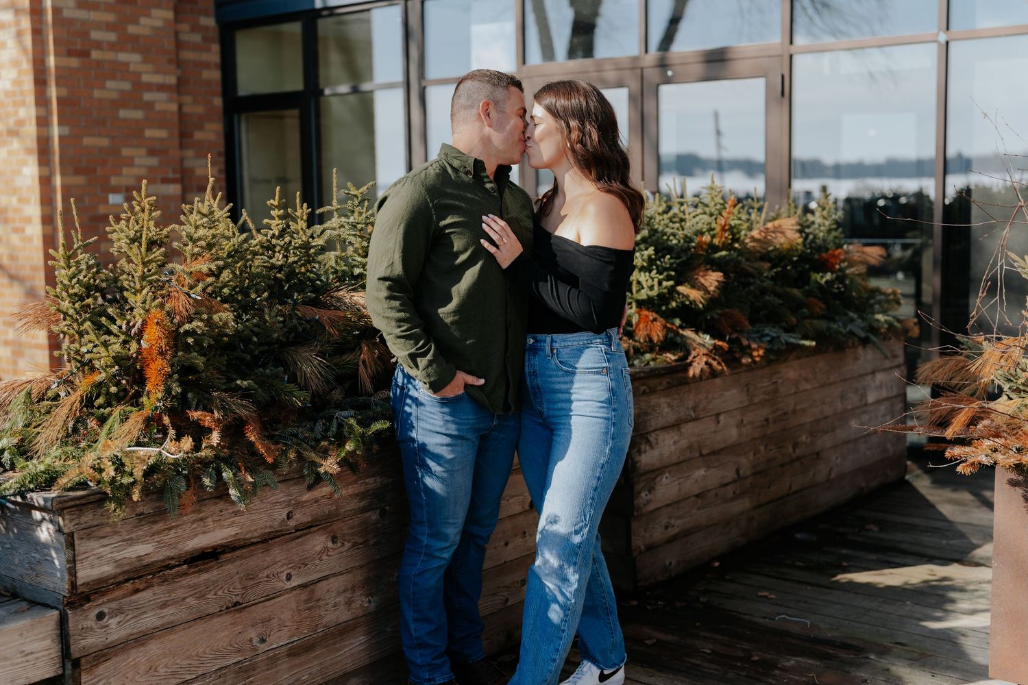 Couple sharing a romantic kiss outdoors near lush greenery planters, wearing casual jeans and dark tops.