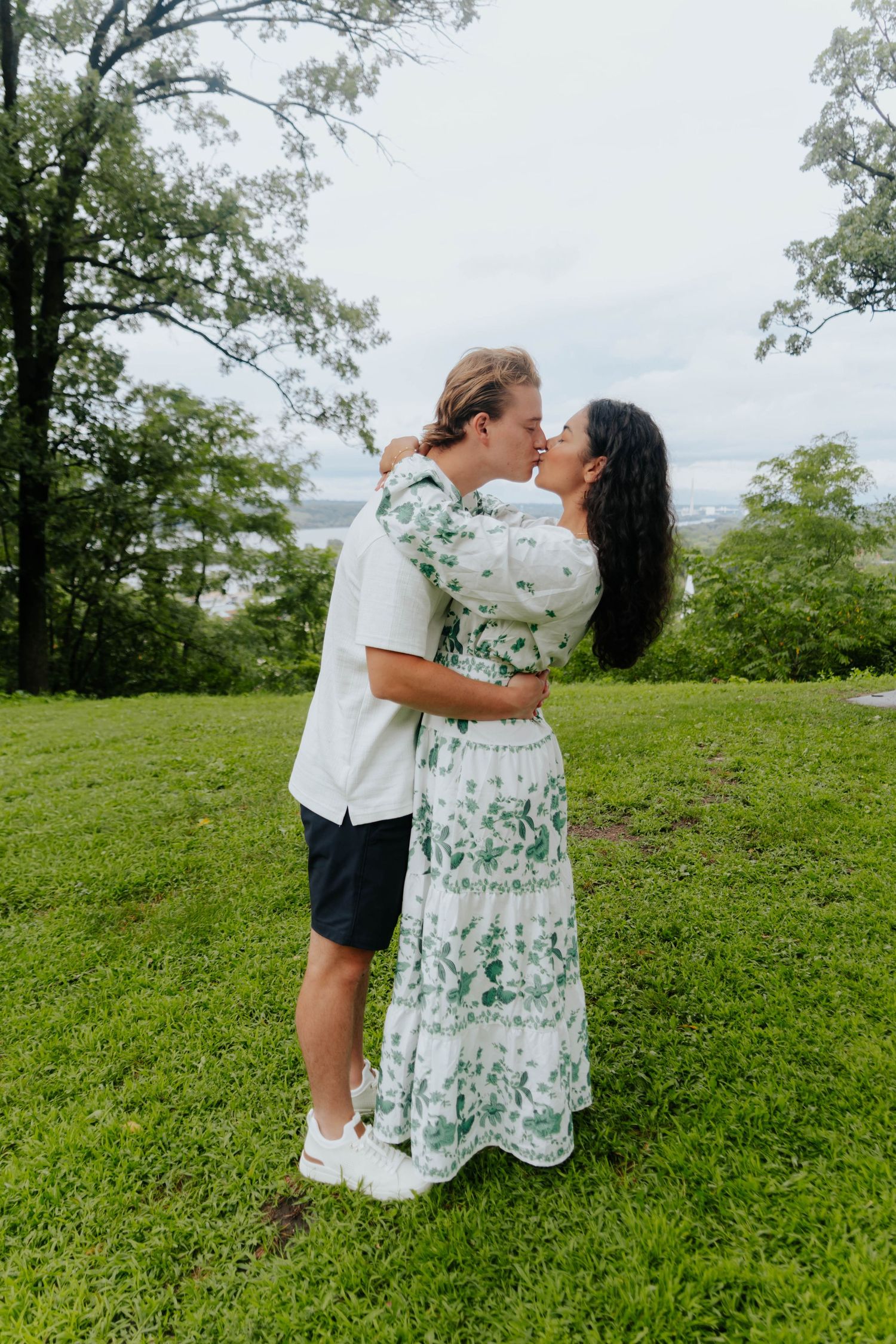 A couple shares a romantic kiss on a lush green lawn surrounded by trees on a cloudy day.
