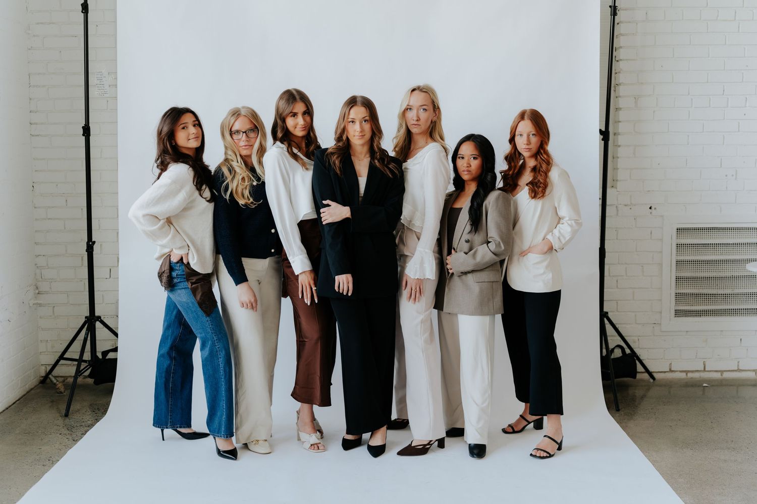 Seven stylish women posing together in elegant black and white outfits against a white studio backdrop.